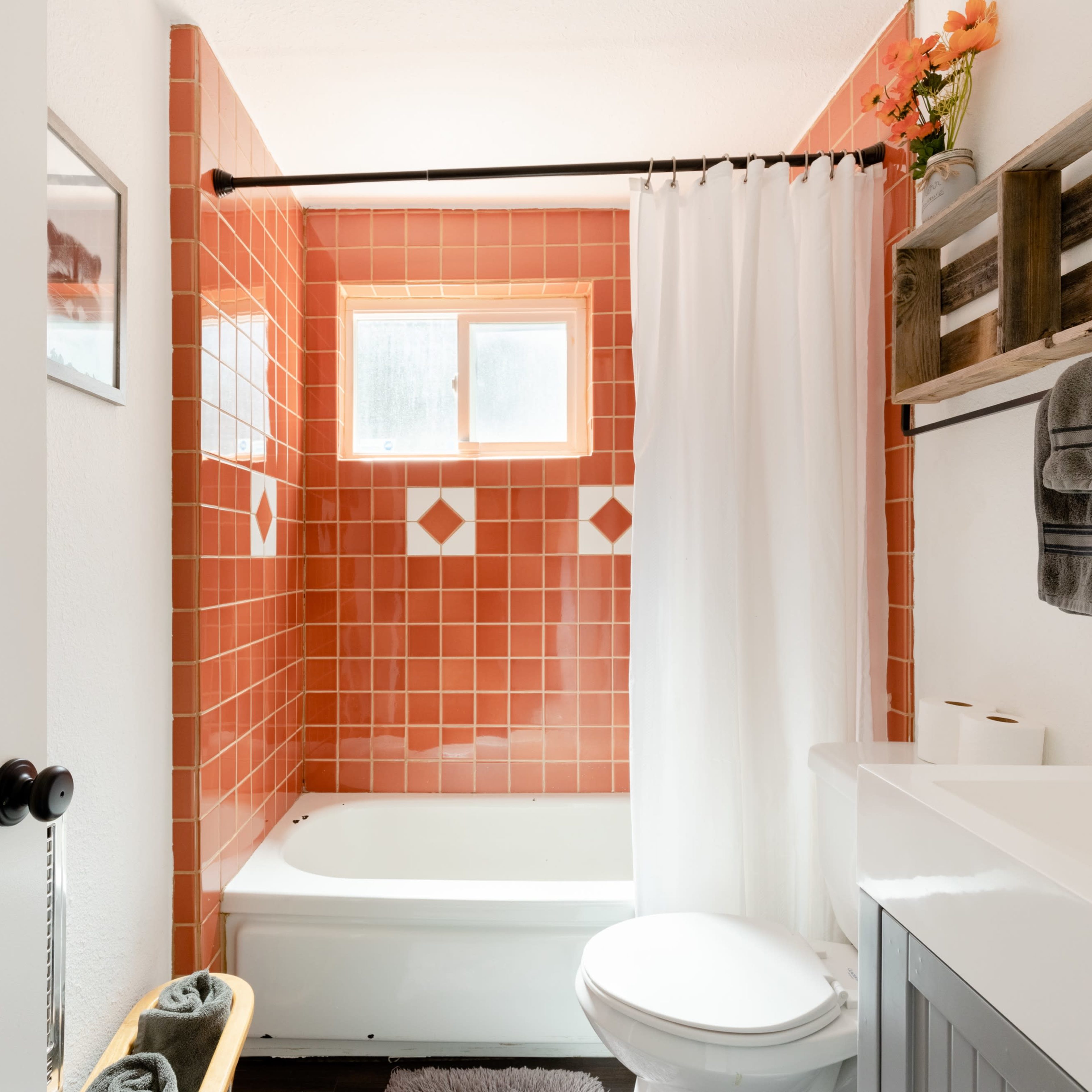 A modern bathroom with a bathtub, orange tiled walls, a shower curtain, and a wooden shelf displaying towels and flowers.