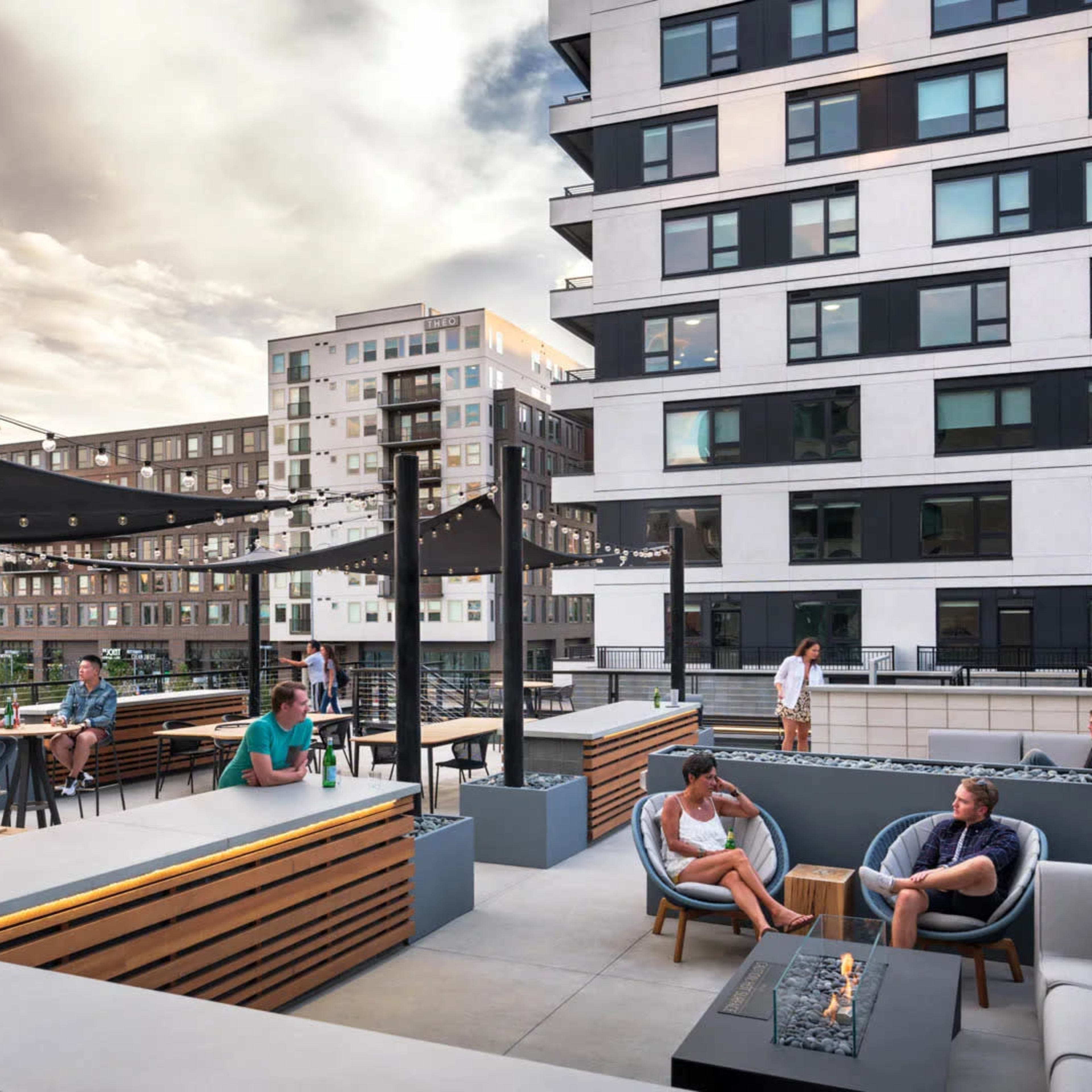 A rooftop terrace features several seating areas, people socializing, and decorative string lights against a backdrop of modern apartment buildings.