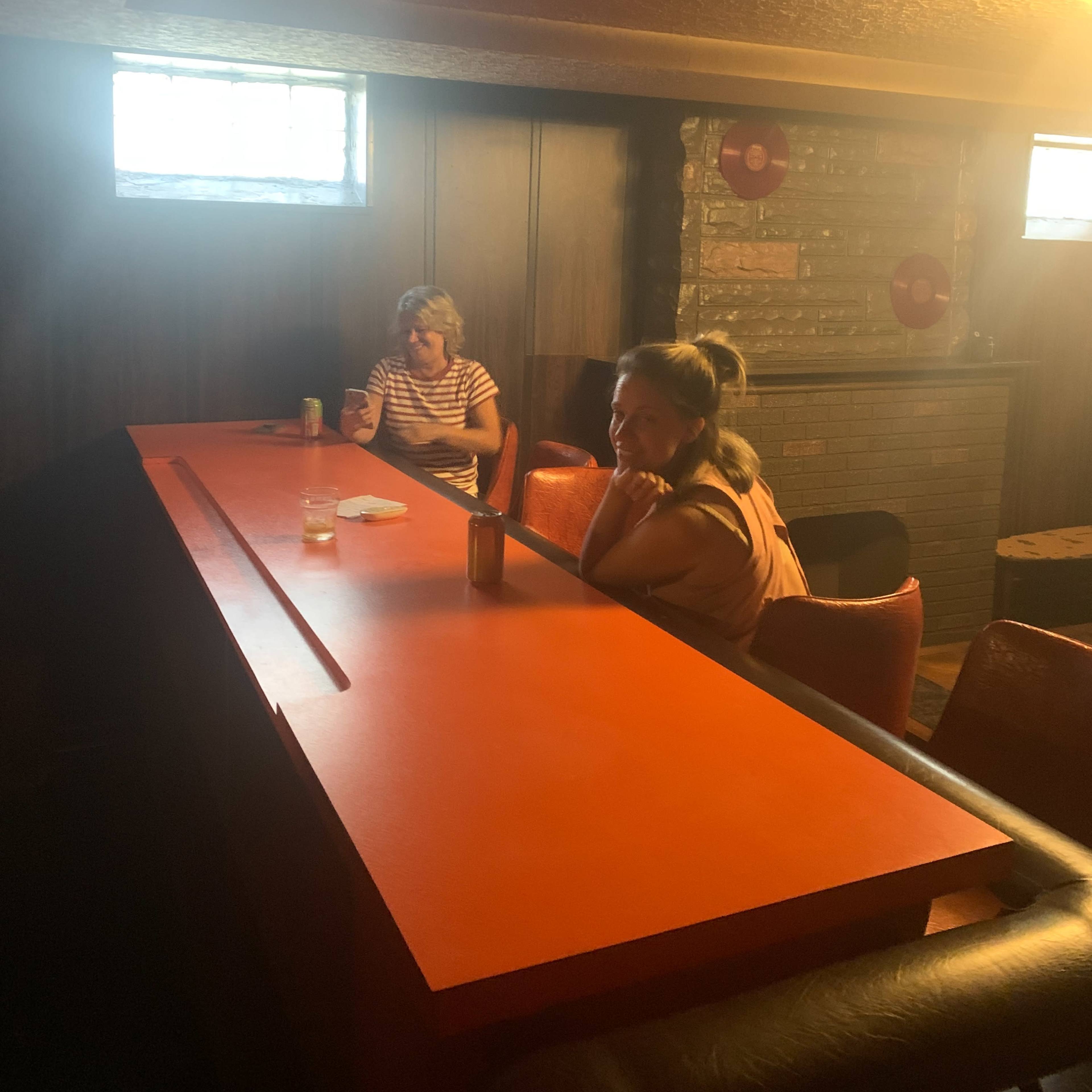 Two people sit at a long orange bar in a dimly lit basement, with a stone fireplace in the background.