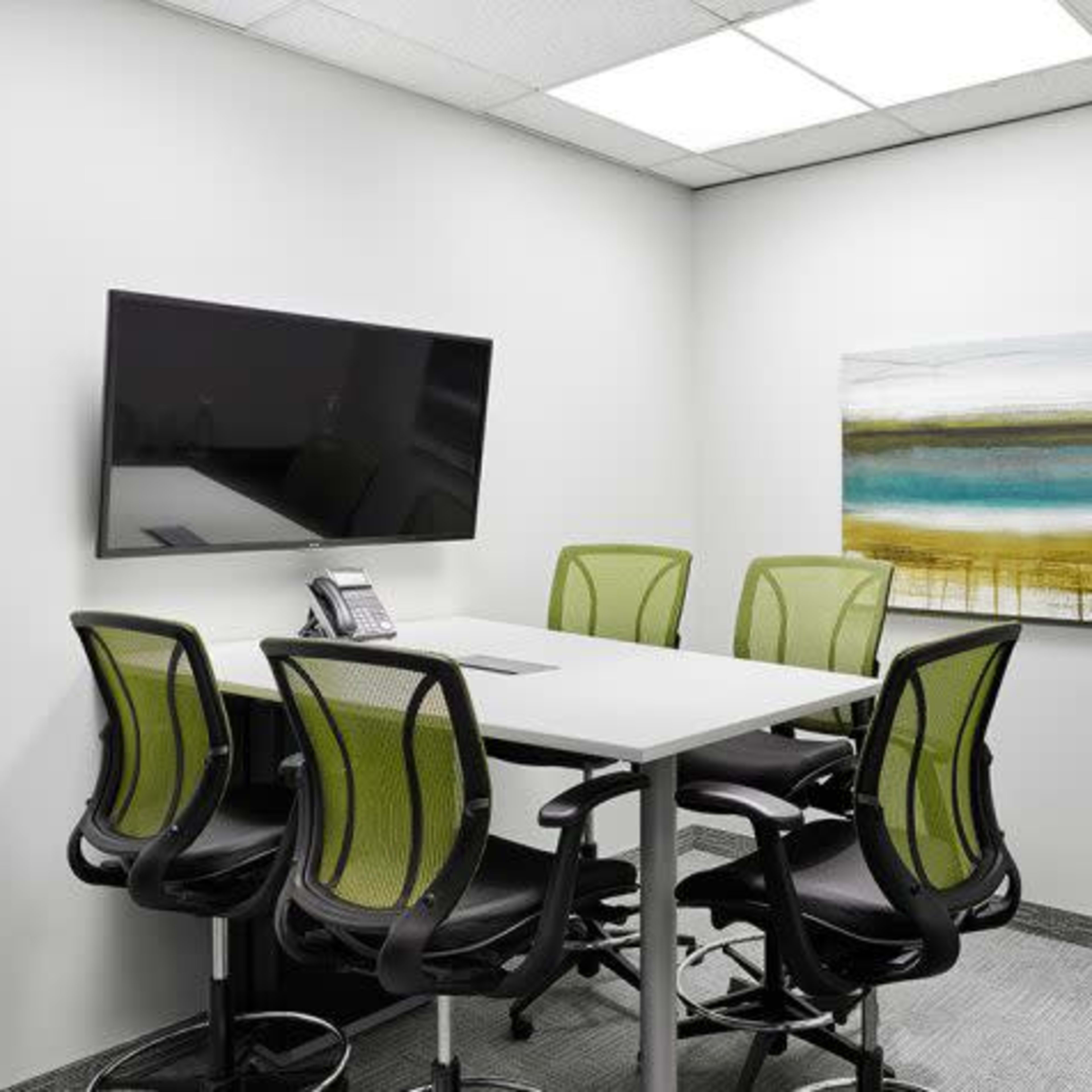 A small conference room with a table surrounded by five green office chairs, a wall-mounted TV, and an abstract painting.
