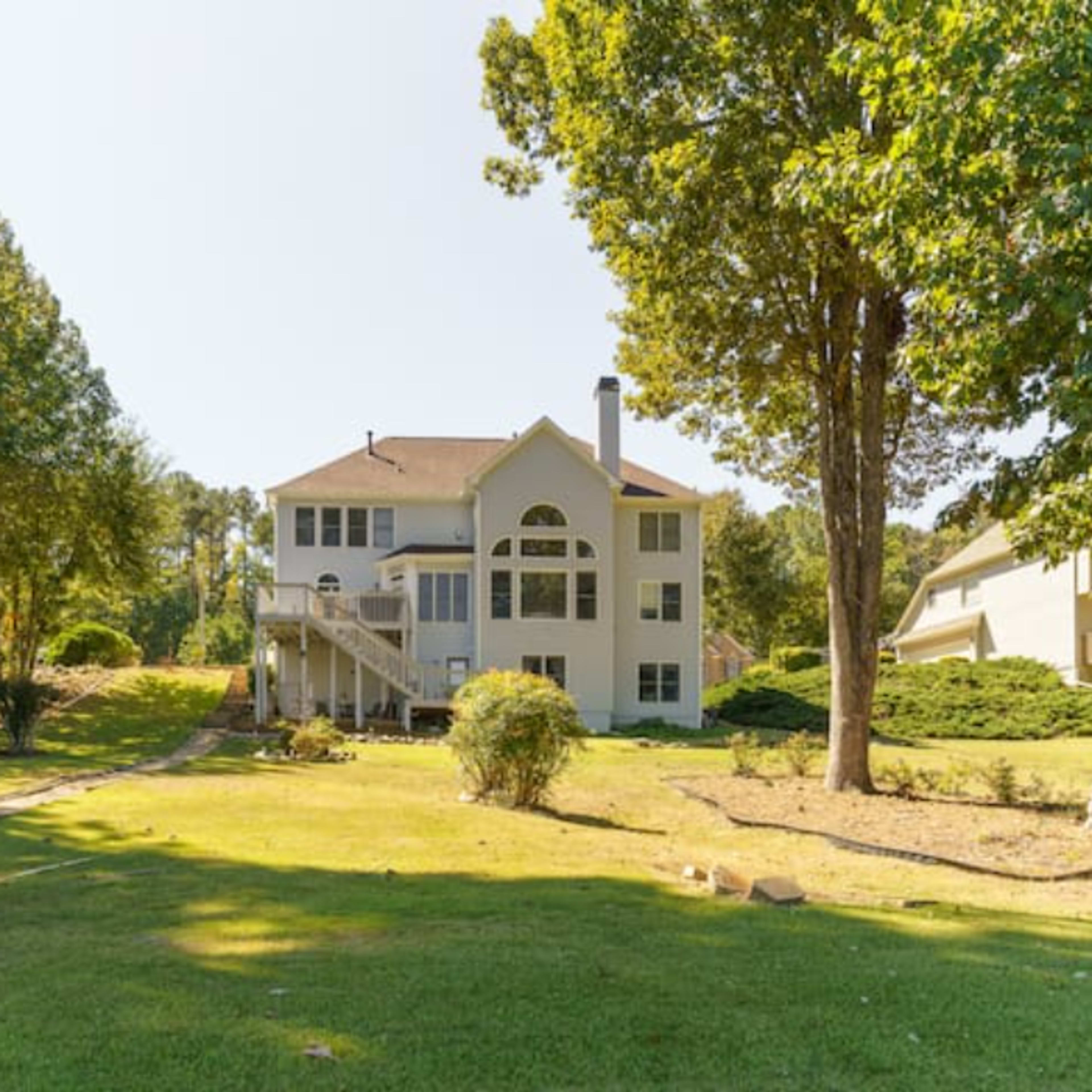 A two-story house with a large deck overlooks a grassy yard surrounded by trees.