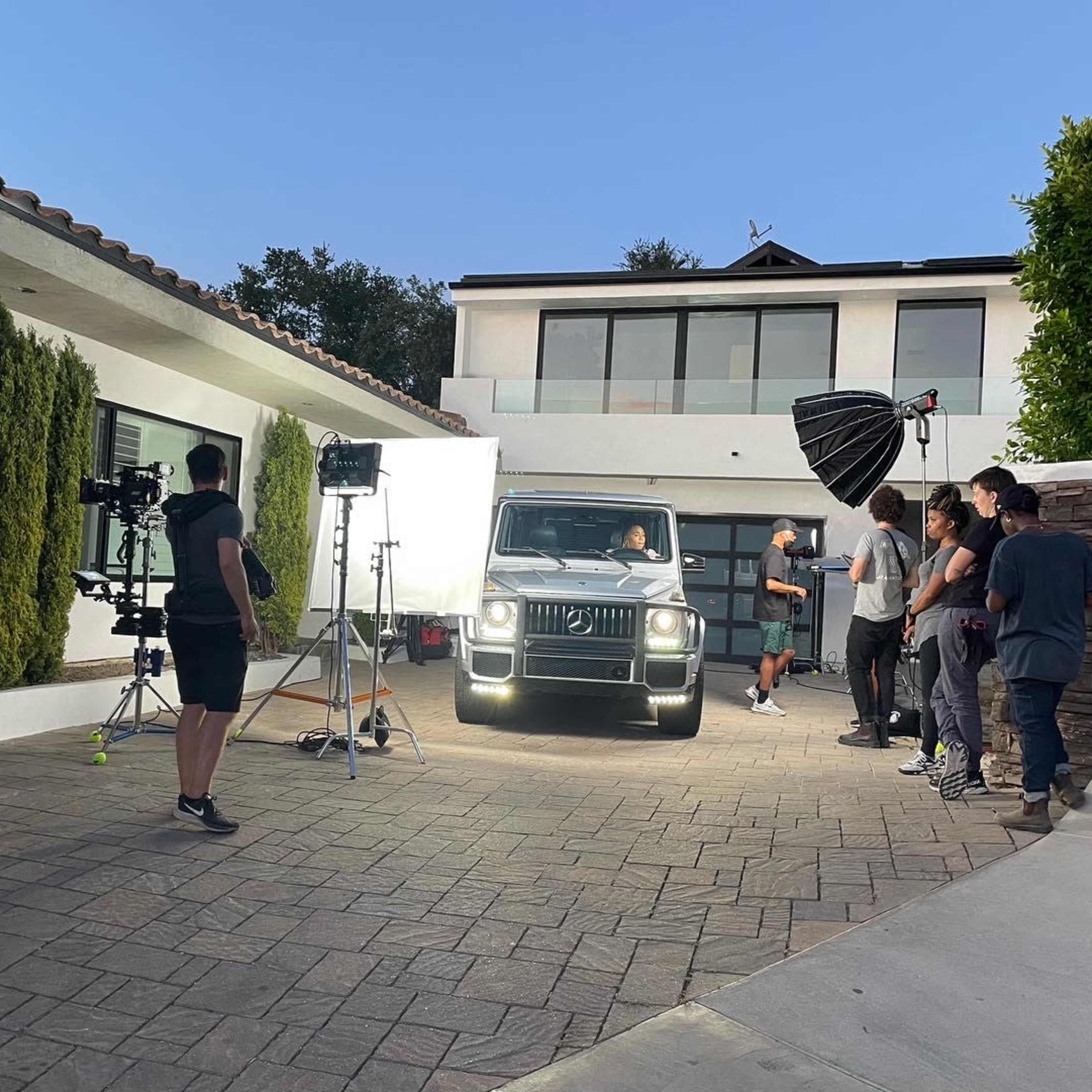 A film crew sets up equipment around a luxury SUV parked in a residential driveway.
