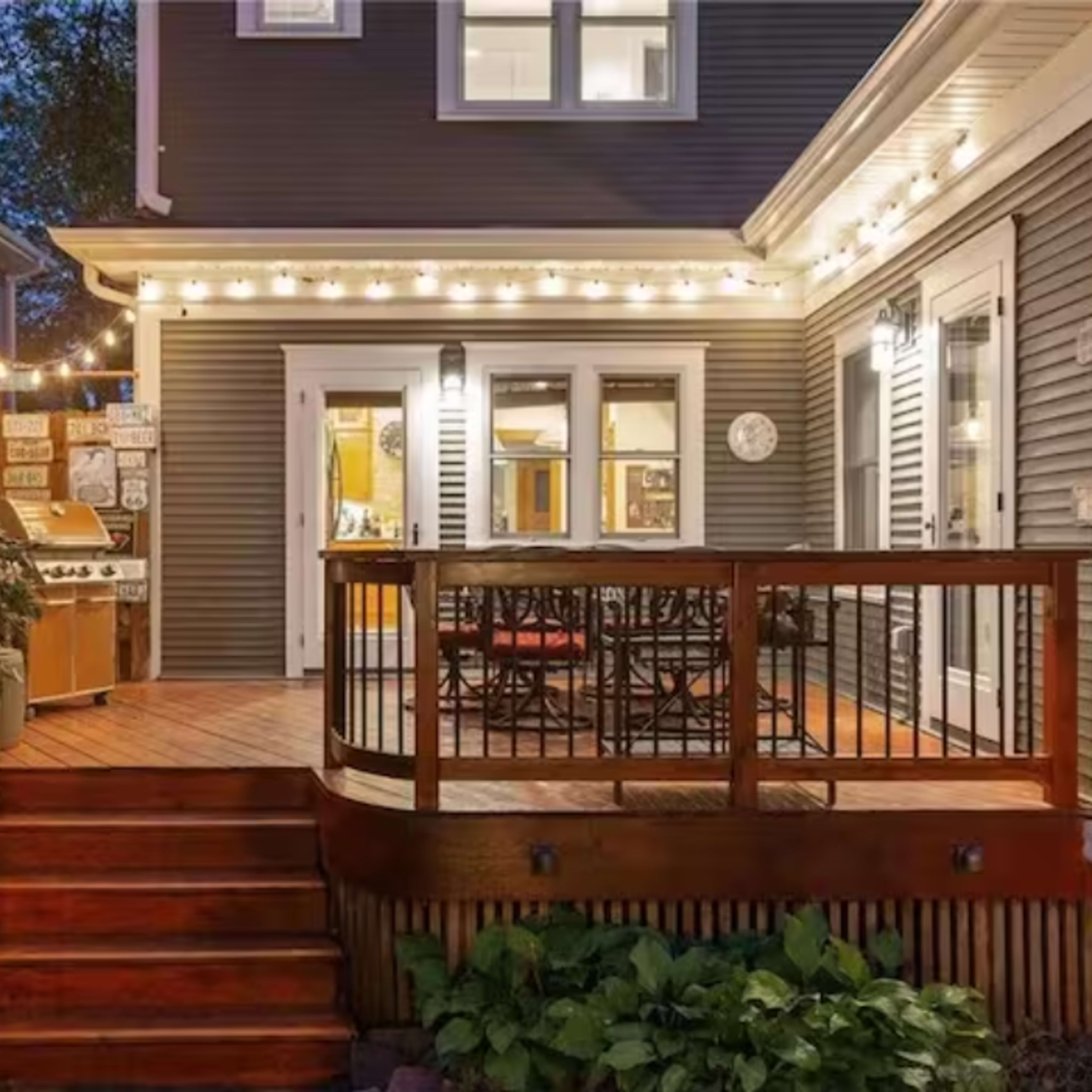 A wooden deck with a railing, leading to a backyard area featuring outdoor seating and string lights, adjacent to a house.