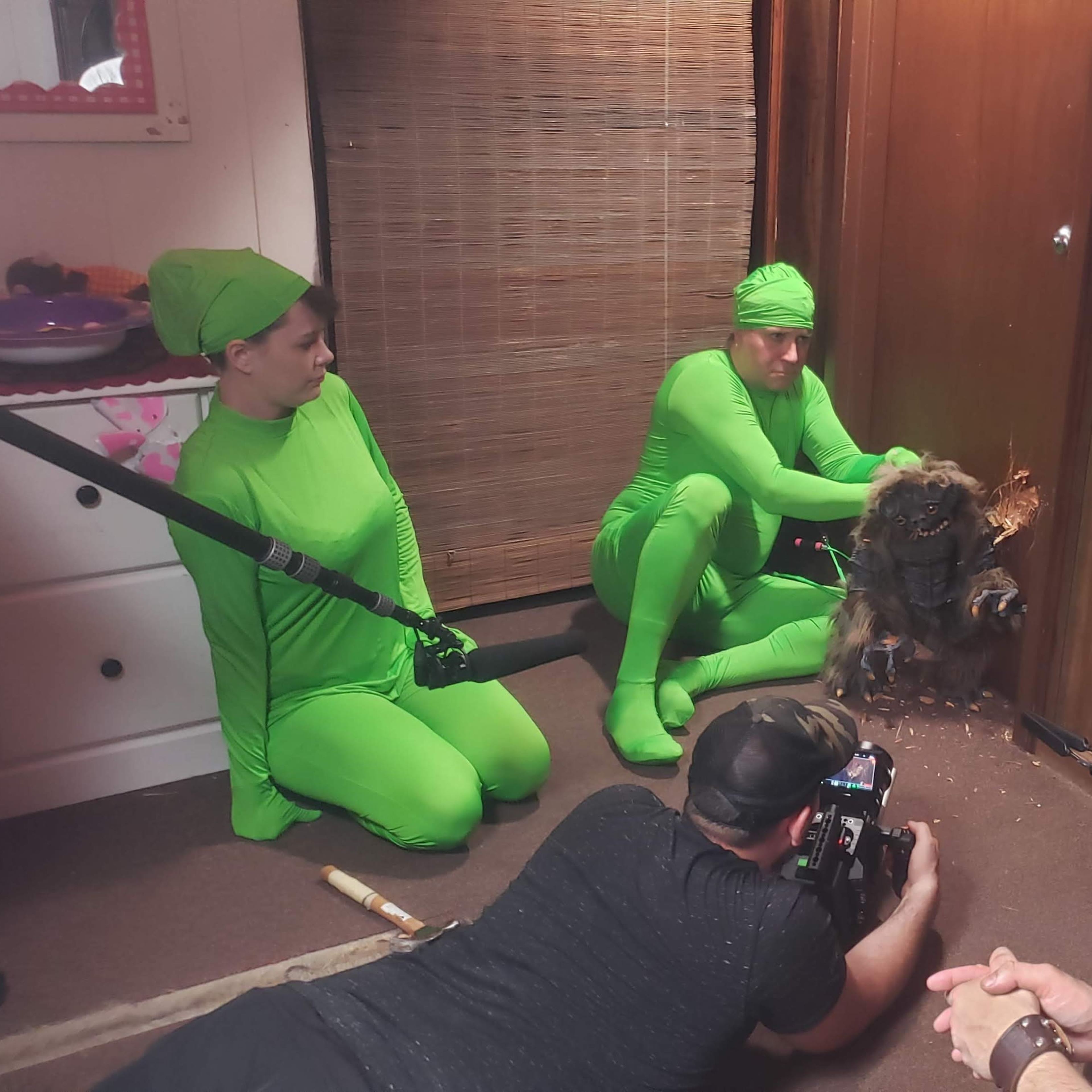 A group of people in green costumes, including one holding a camera, is filming a scene with a dog near a wooden door.