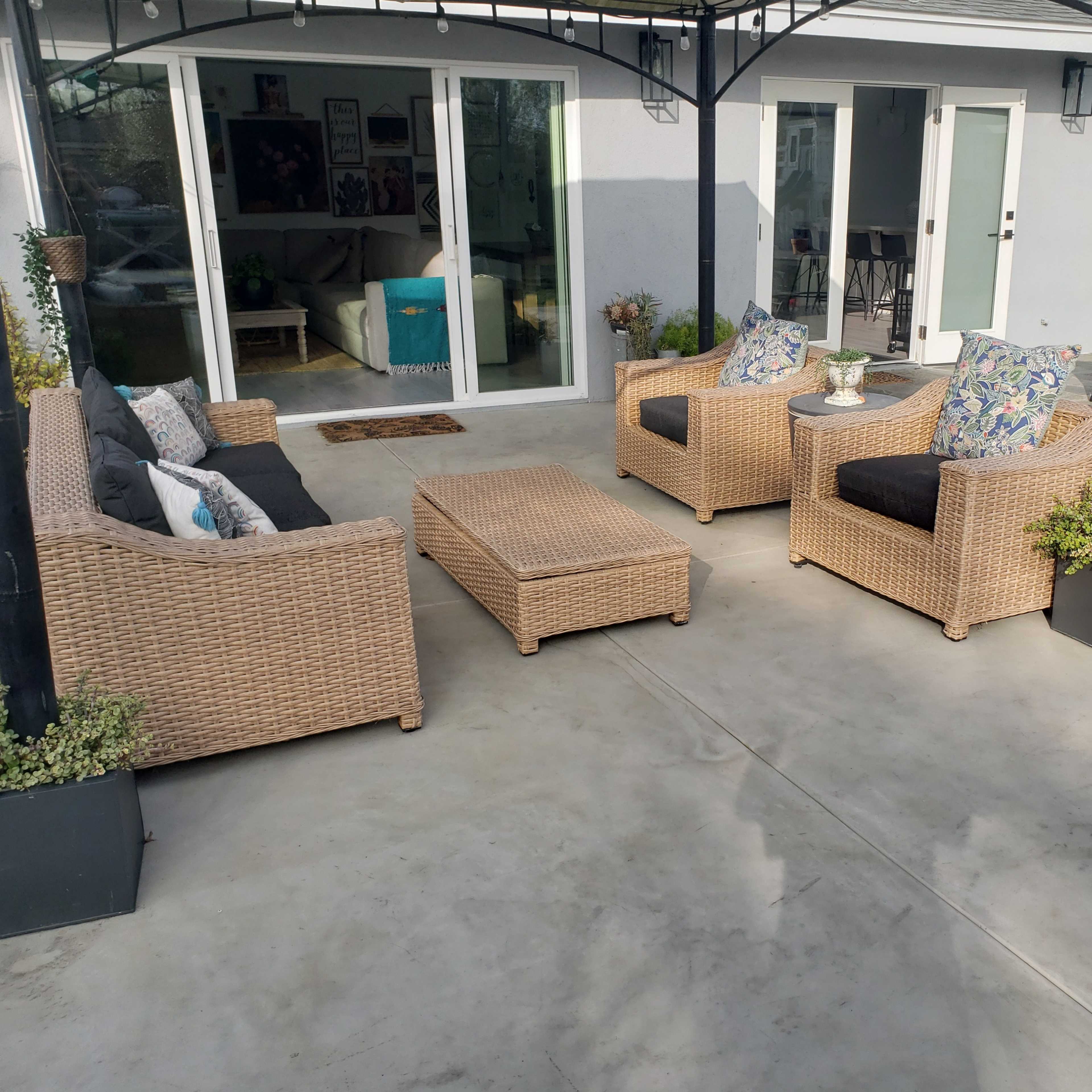 A patio area features four wicker chairs with cushions, a matching coffee table, and potted plants, surrounded by a concrete floor and a sliding glass door.