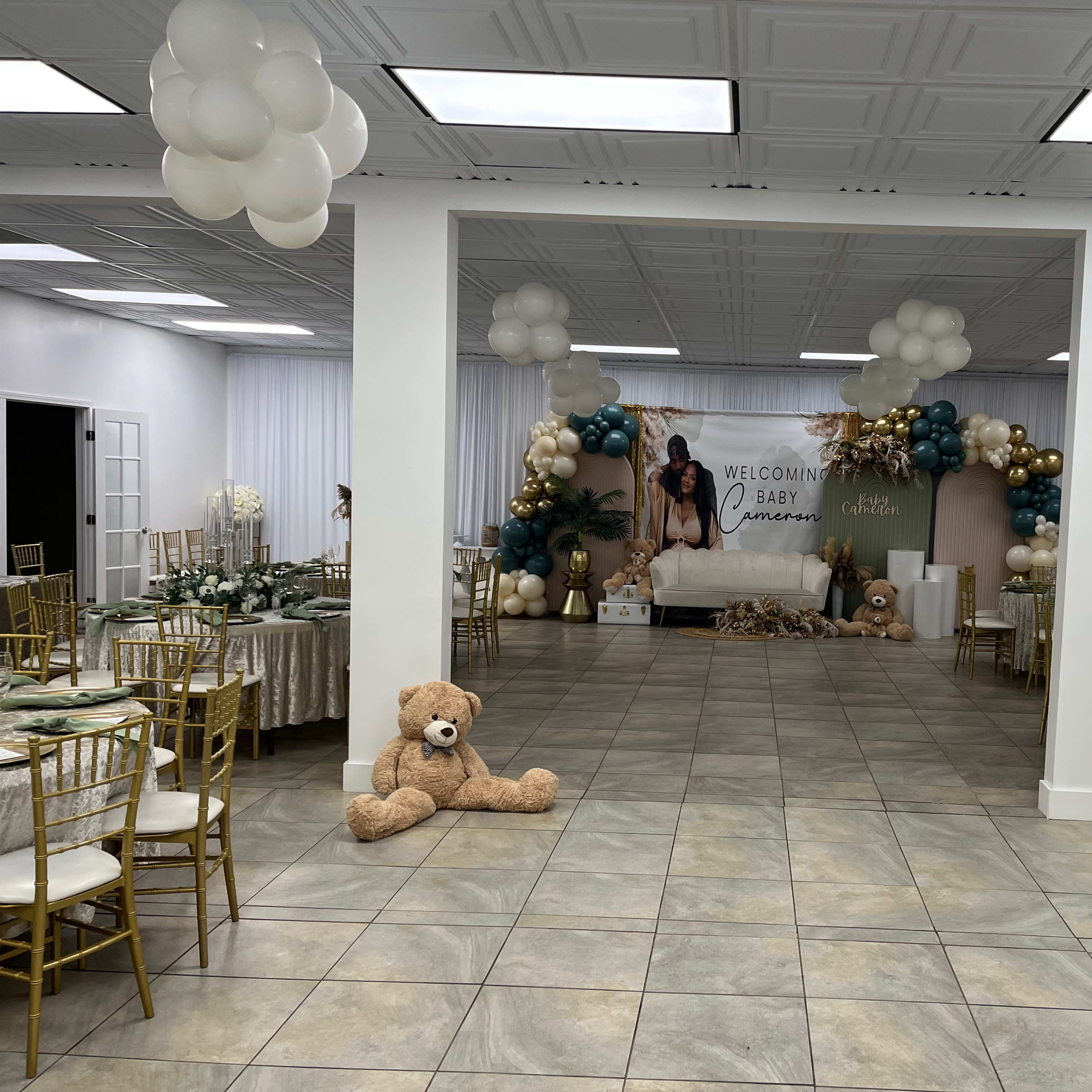 A decorated event space set up for a baby shower, featuring tables with gold chairs, a sofa adorned with balloons, and a large banner welcoming the baby.