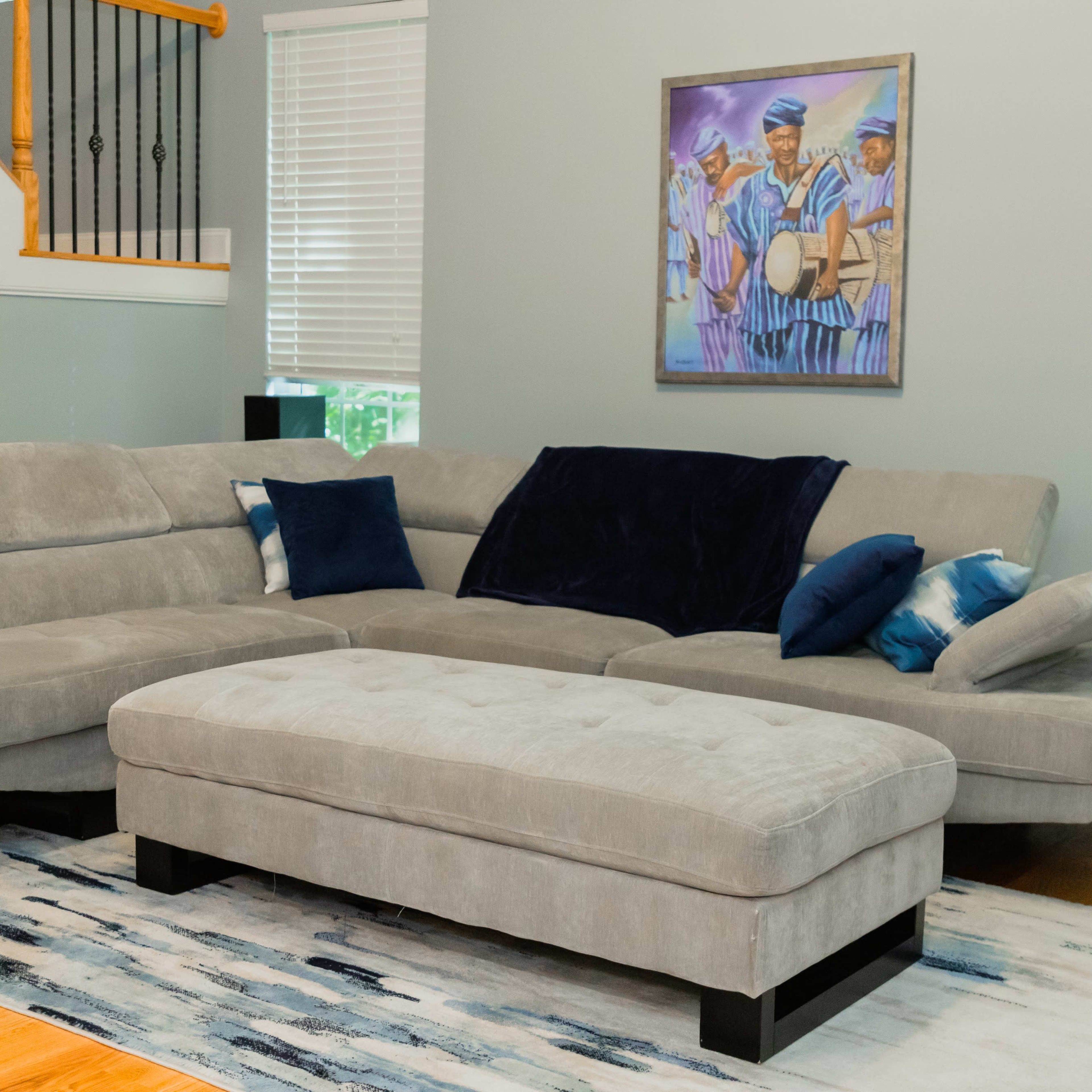 A gray sectional sofa with navy blue and patterned pillows sits on a blue and white area rug in a room with light gray walls and a colorful painting.