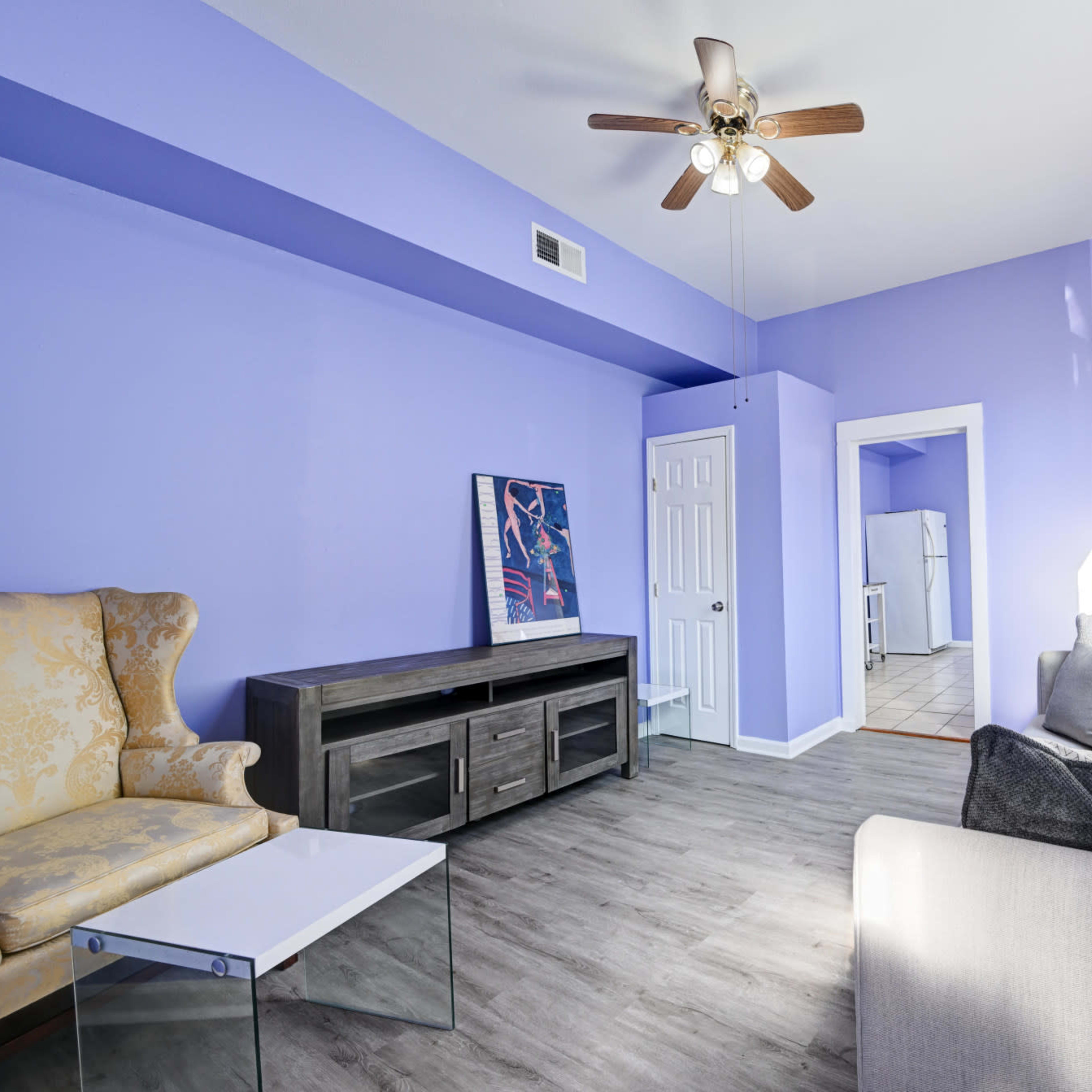 A living room with purple walls, a ceiling fan, and a mix of light furniture including a yellow armchair and a gray sofa.