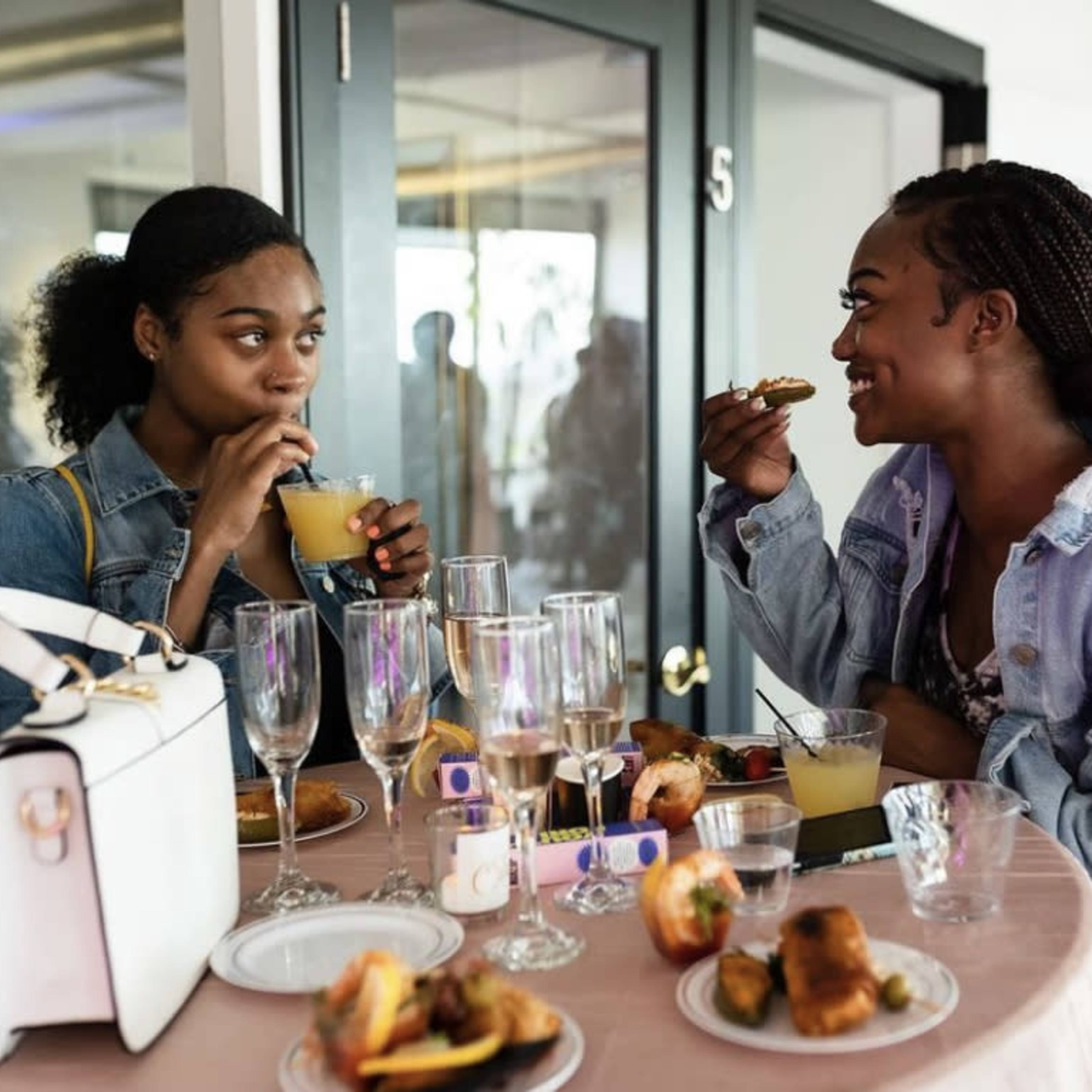 Two women are seated at a table filled with food and drinks, enjoying a conversation while eating and drinking from glasses.