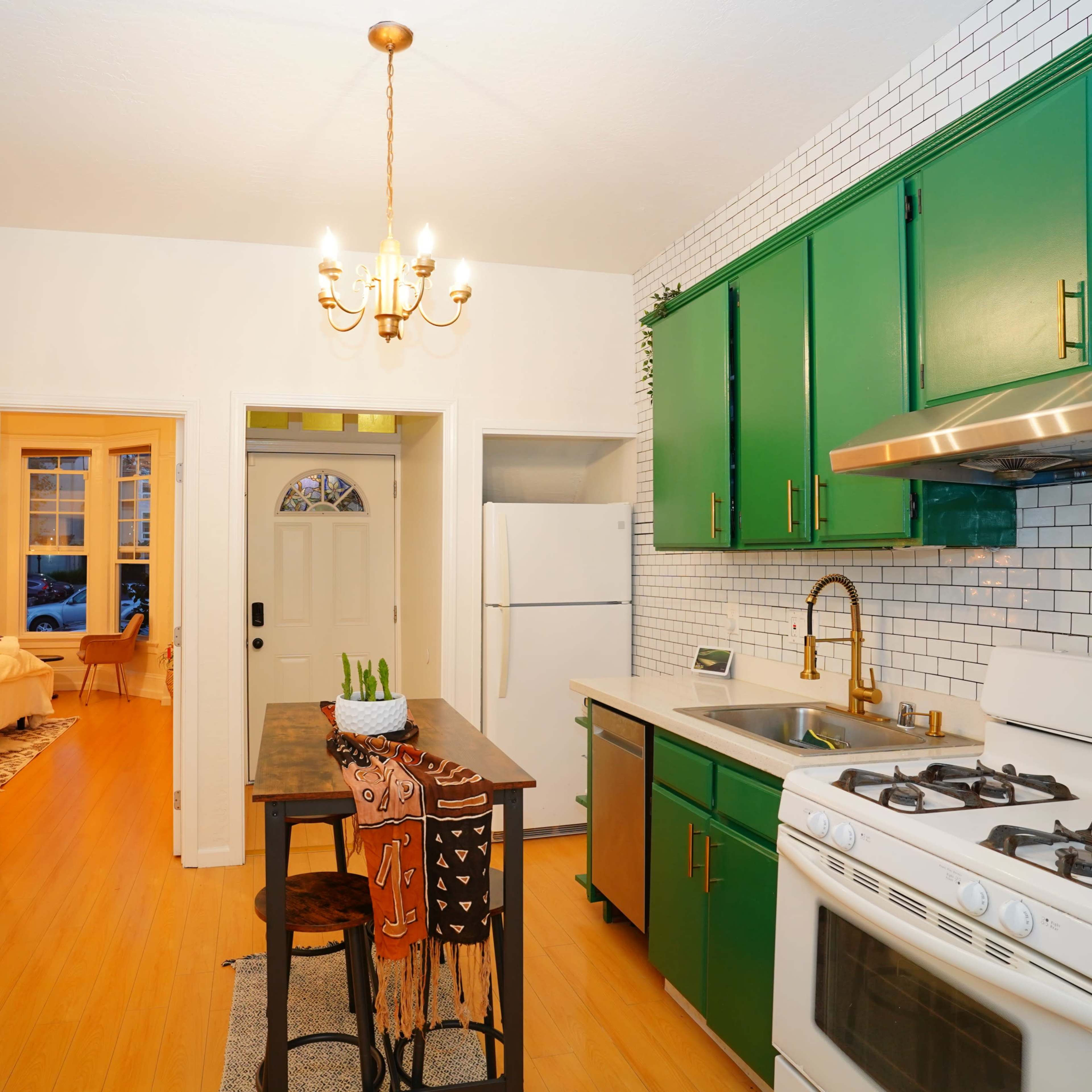 A bright kitchen features green cabinets, white tiled walls, and a small dining table with two chairs, with an entrance leading to another room.