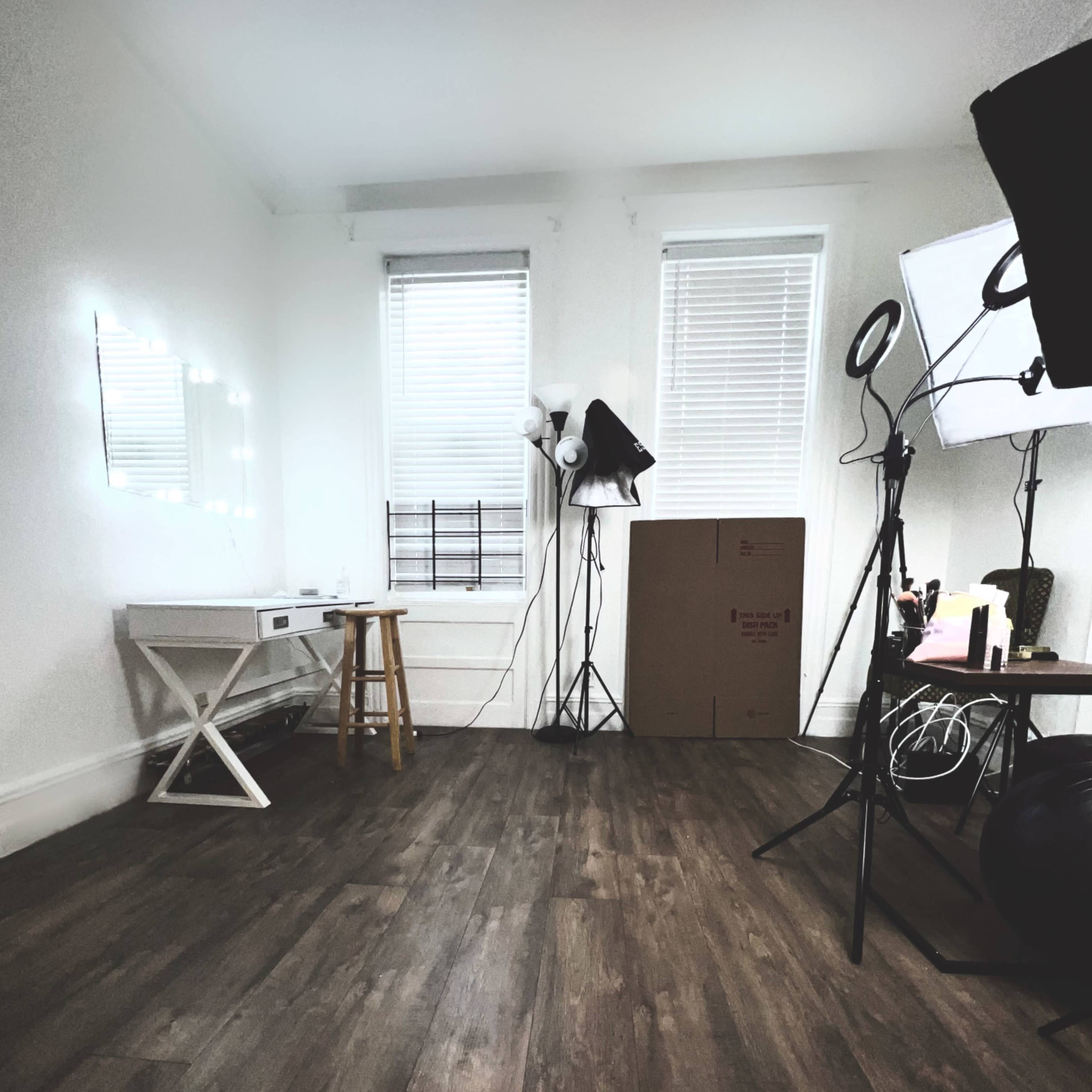 A well-lit room features a white table with a stool, a cardboard box, a chair, and studio lighting equipment against the walls.
