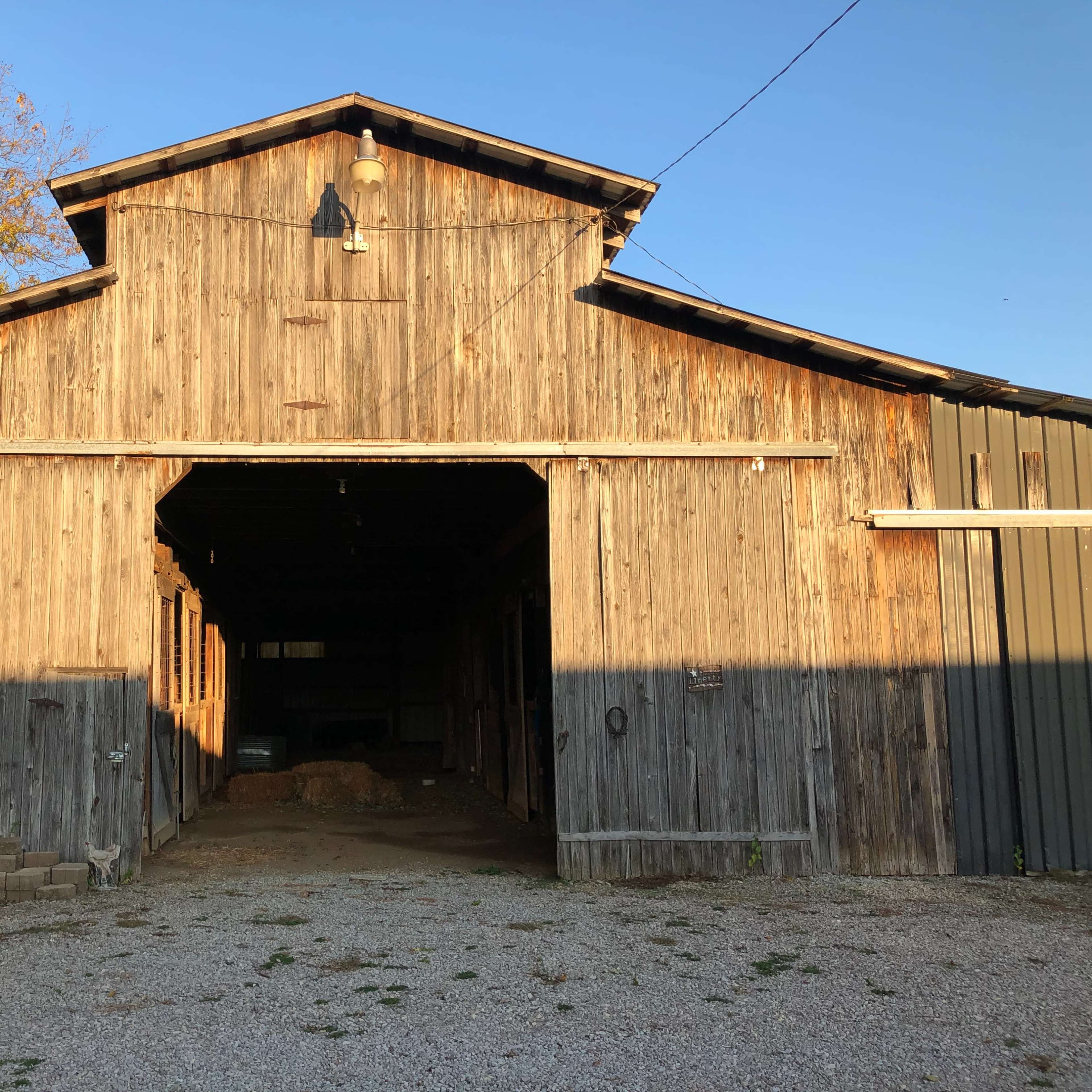 A weathered wooden barn with an open entrance sits on a gravel path under a clear blue sky.