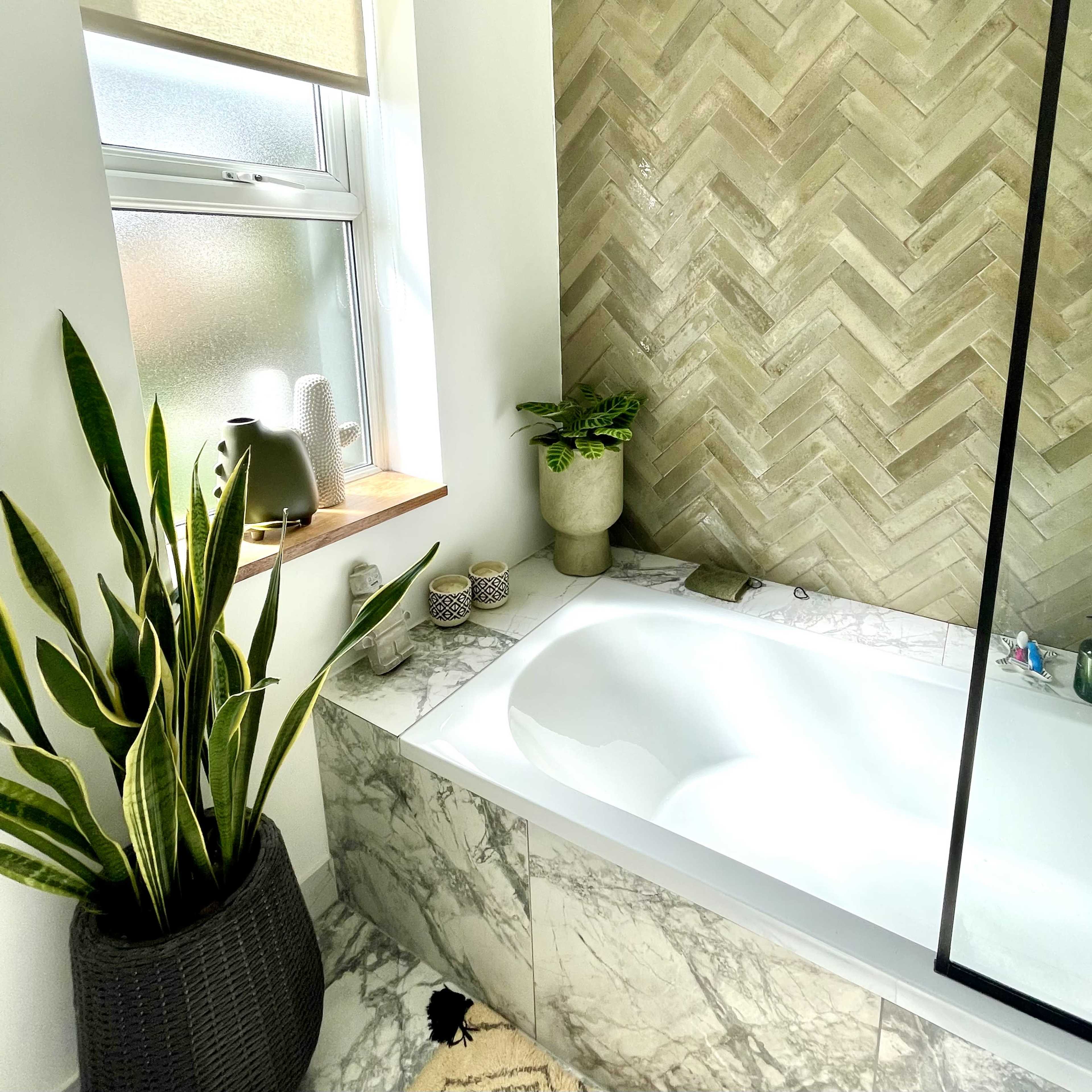 A modern bathroom with a herringbone-patterned tiled wall, a white bathtub, and a plant beside it.