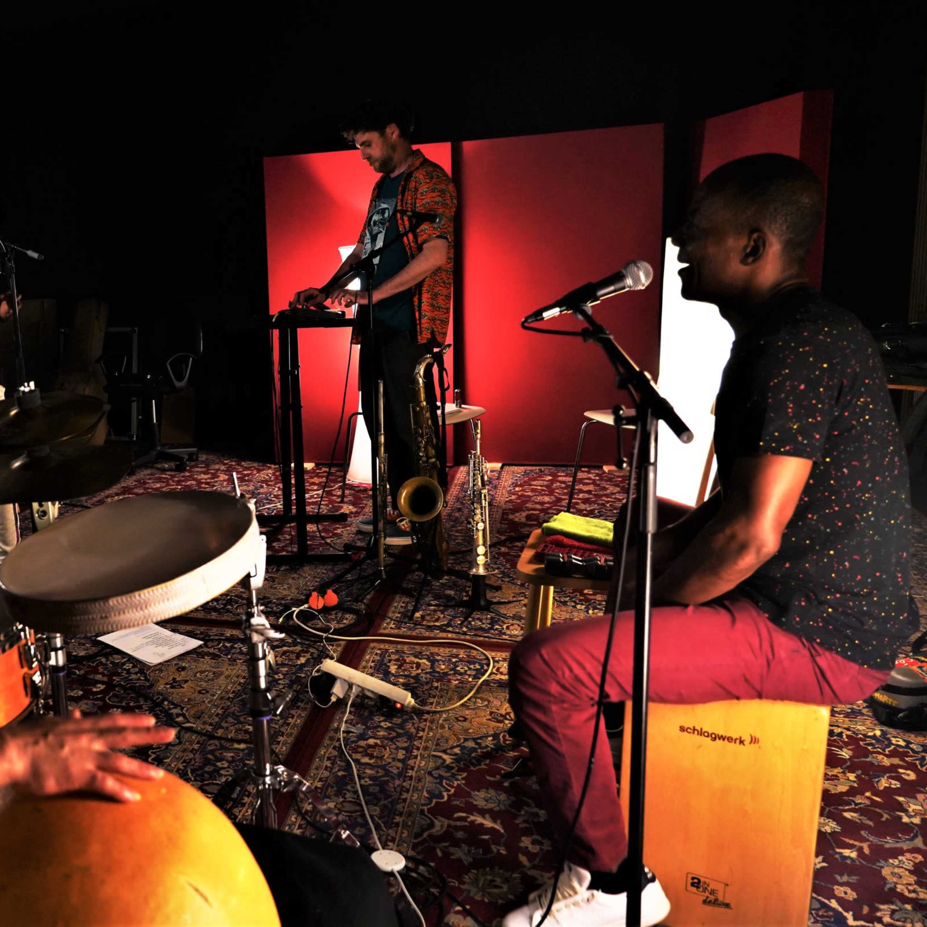 A band performs in a studio, featuring a drummer, a saxophonist, and two musicians at microphones.