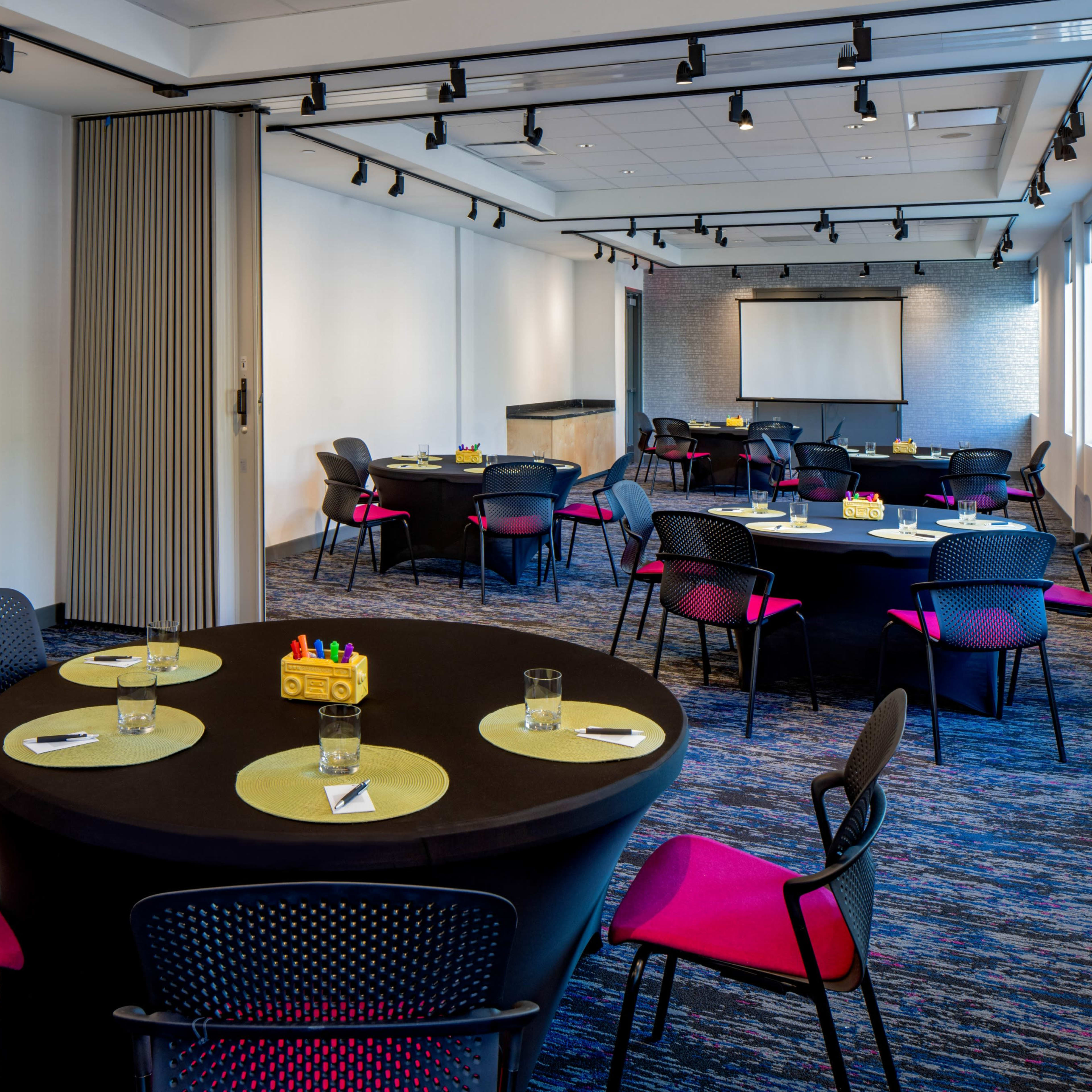 A modern conference room features round tables with black linens and pink chairs, arranged for a meeting with a projector screen at the front.