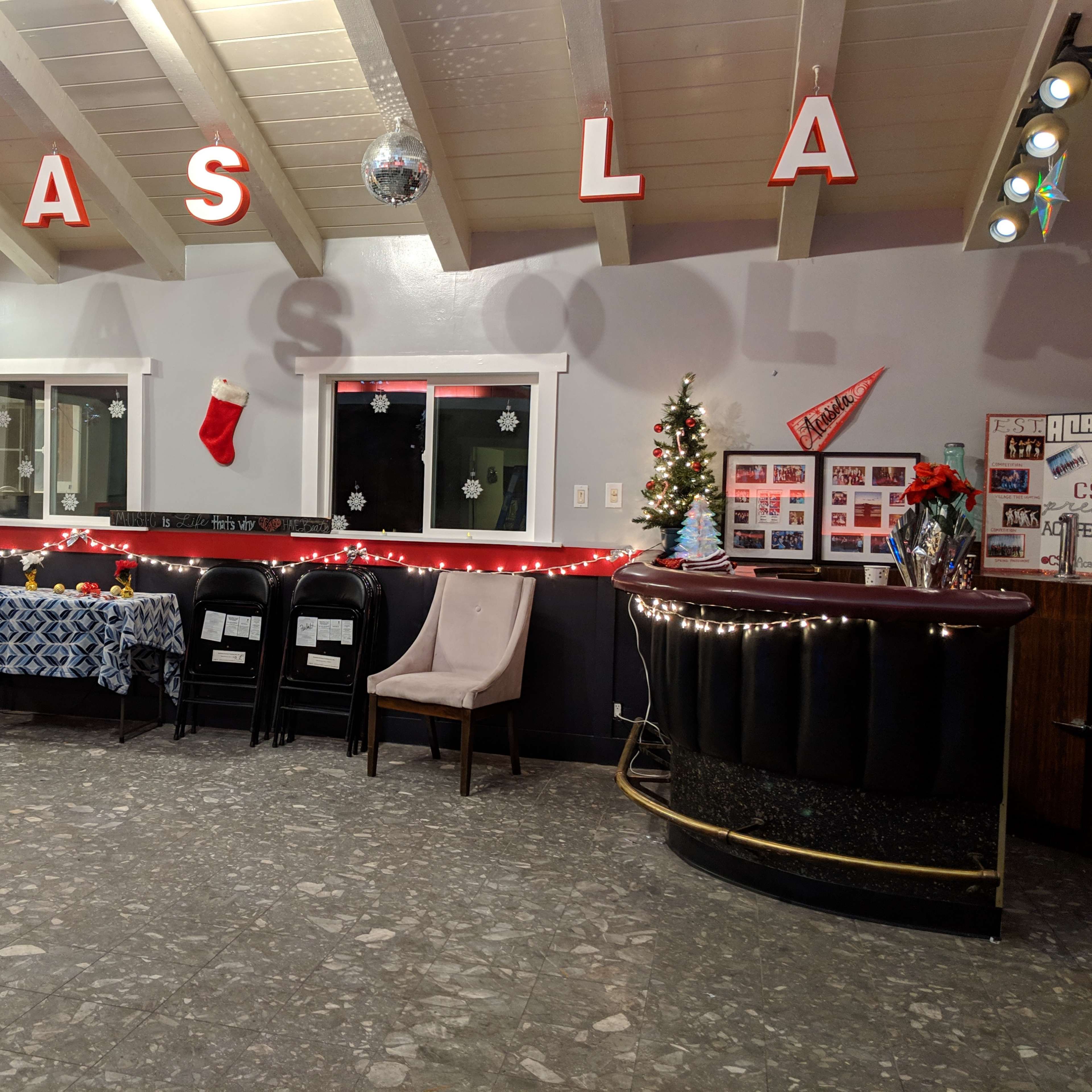 The interior of a warmly lit room features a decorated bar, a table covered with a blue cloth, black chairs, and festive decorations, including a Christmas stocking.