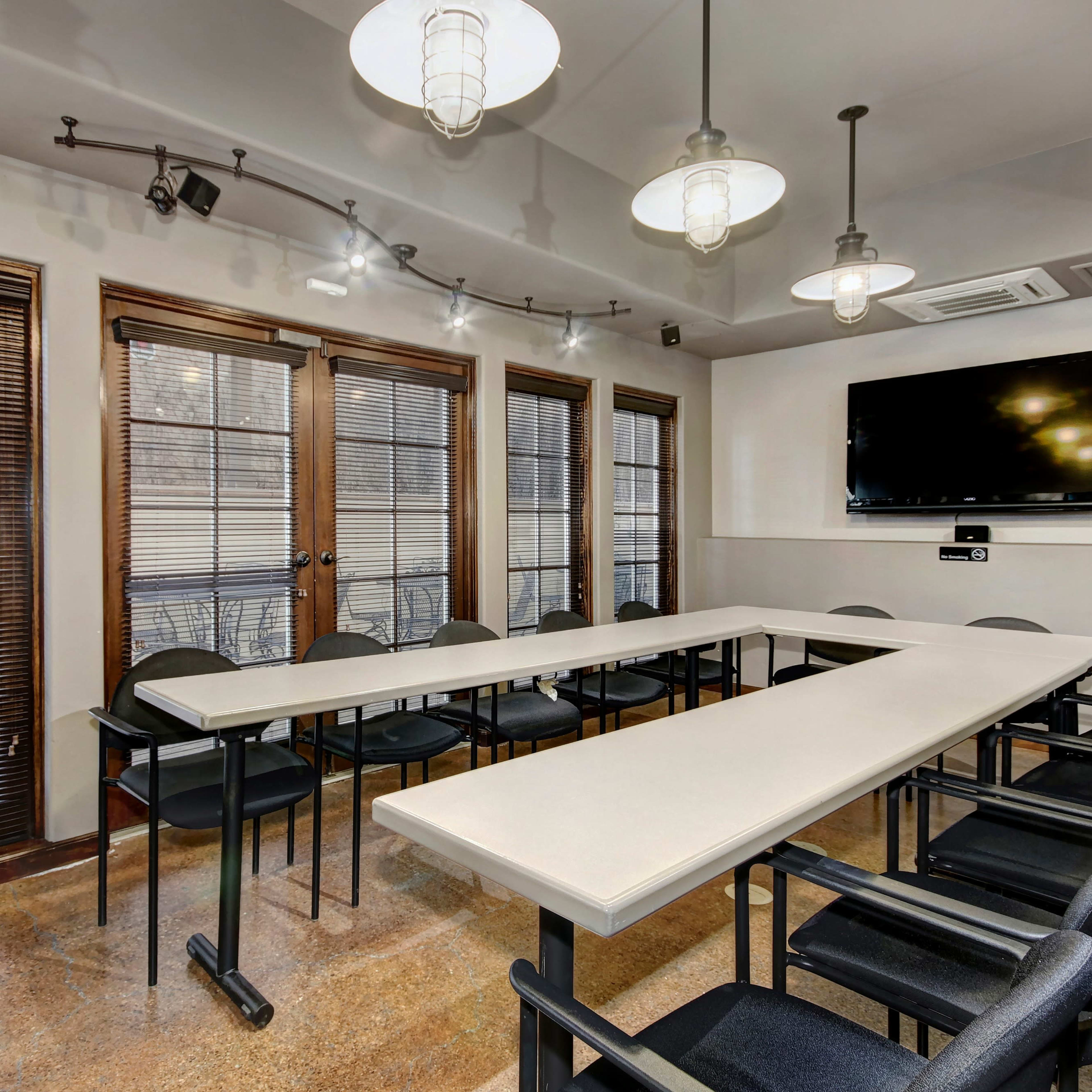 A conference room is set up with a long table surrounded by chairs, and large windows allowing natural light, equipped with a television on the wall.