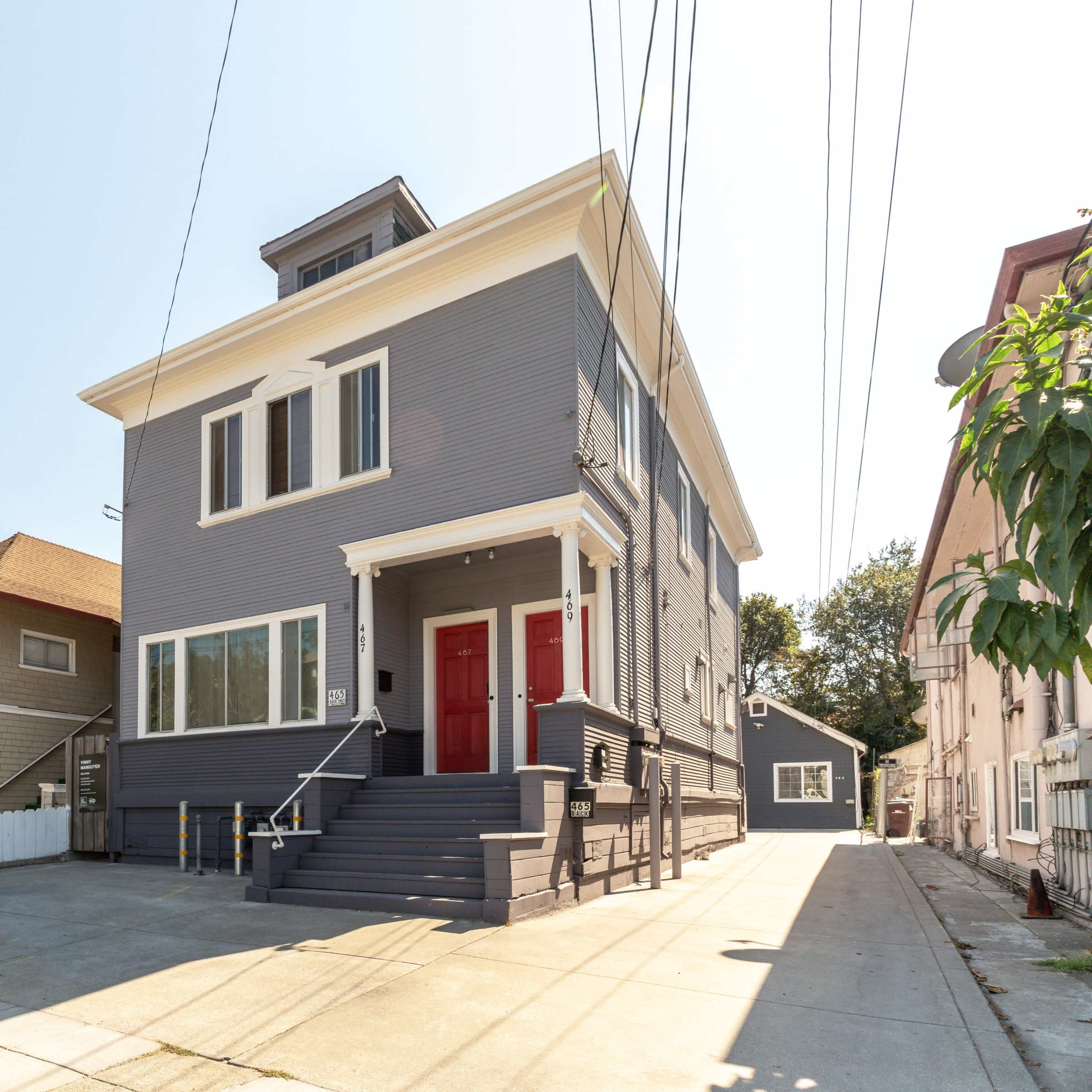 A two-story house with gray siding and red doors, located on a street lined with other residential buildings.