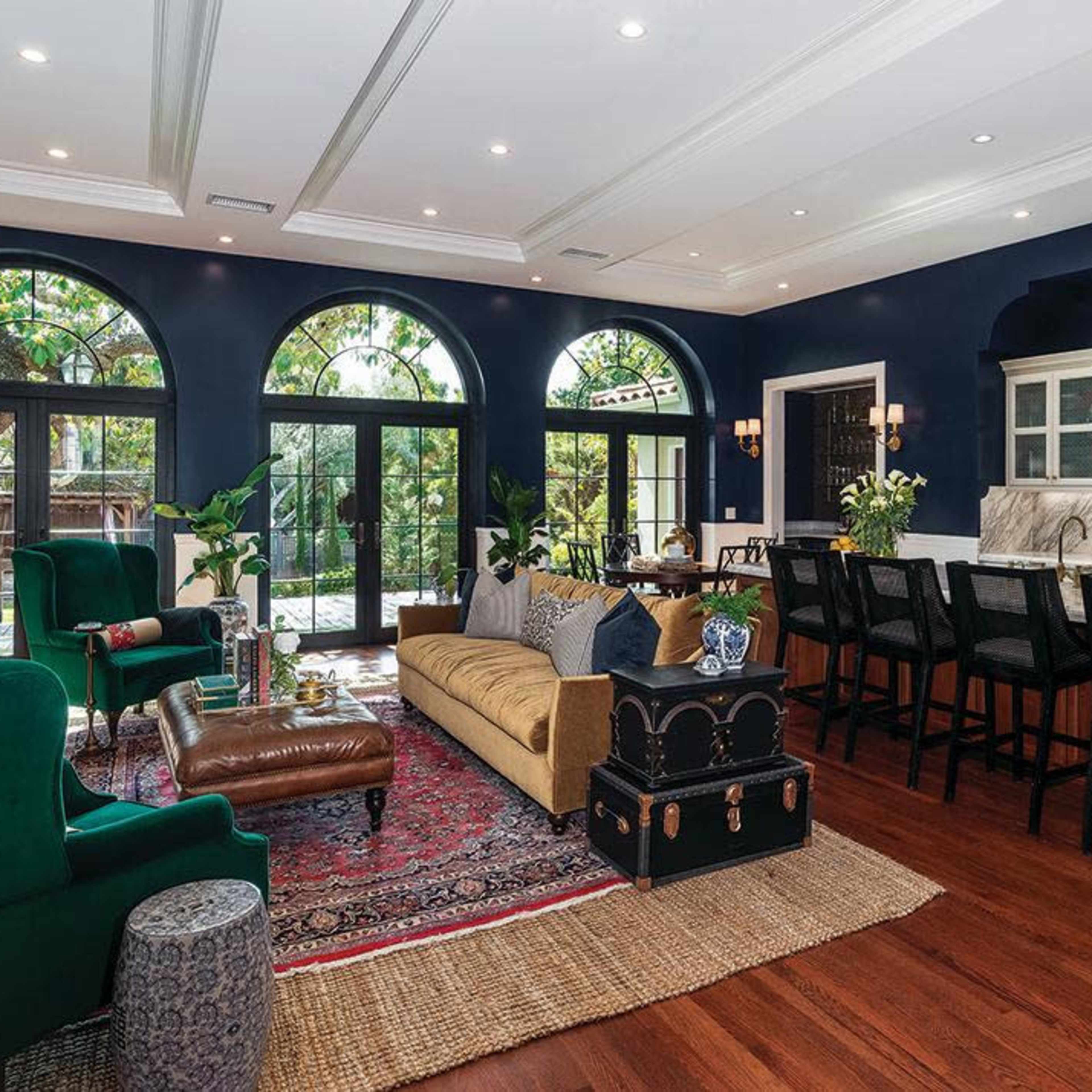 A spacious living area features a dark blue color scheme, large arched windows, and a mix of elegant furniture including a green velvet chair and a patterned area rug.