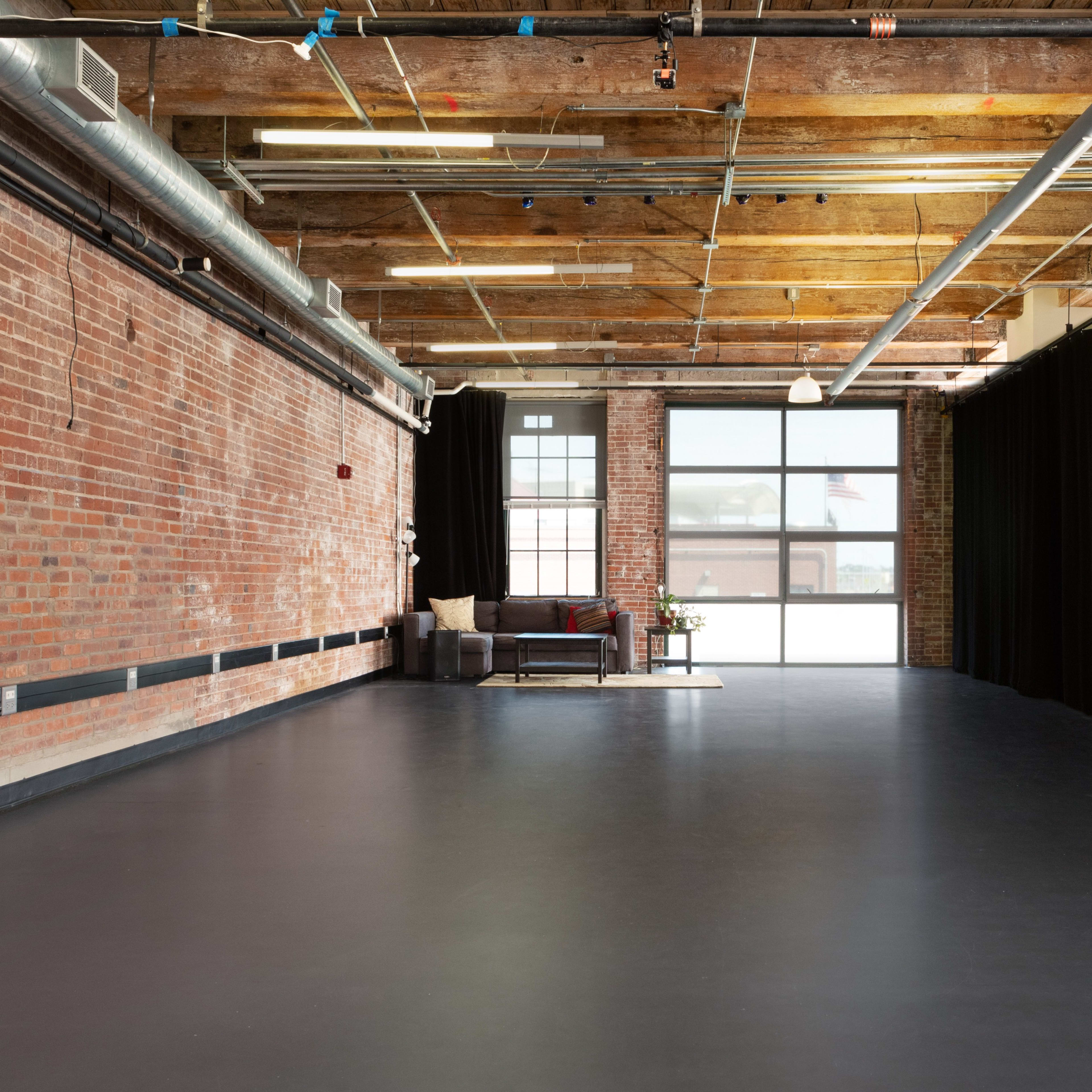 An industrial-style interior space with exposed brick walls, a large window, and a small seating area near a curtain.