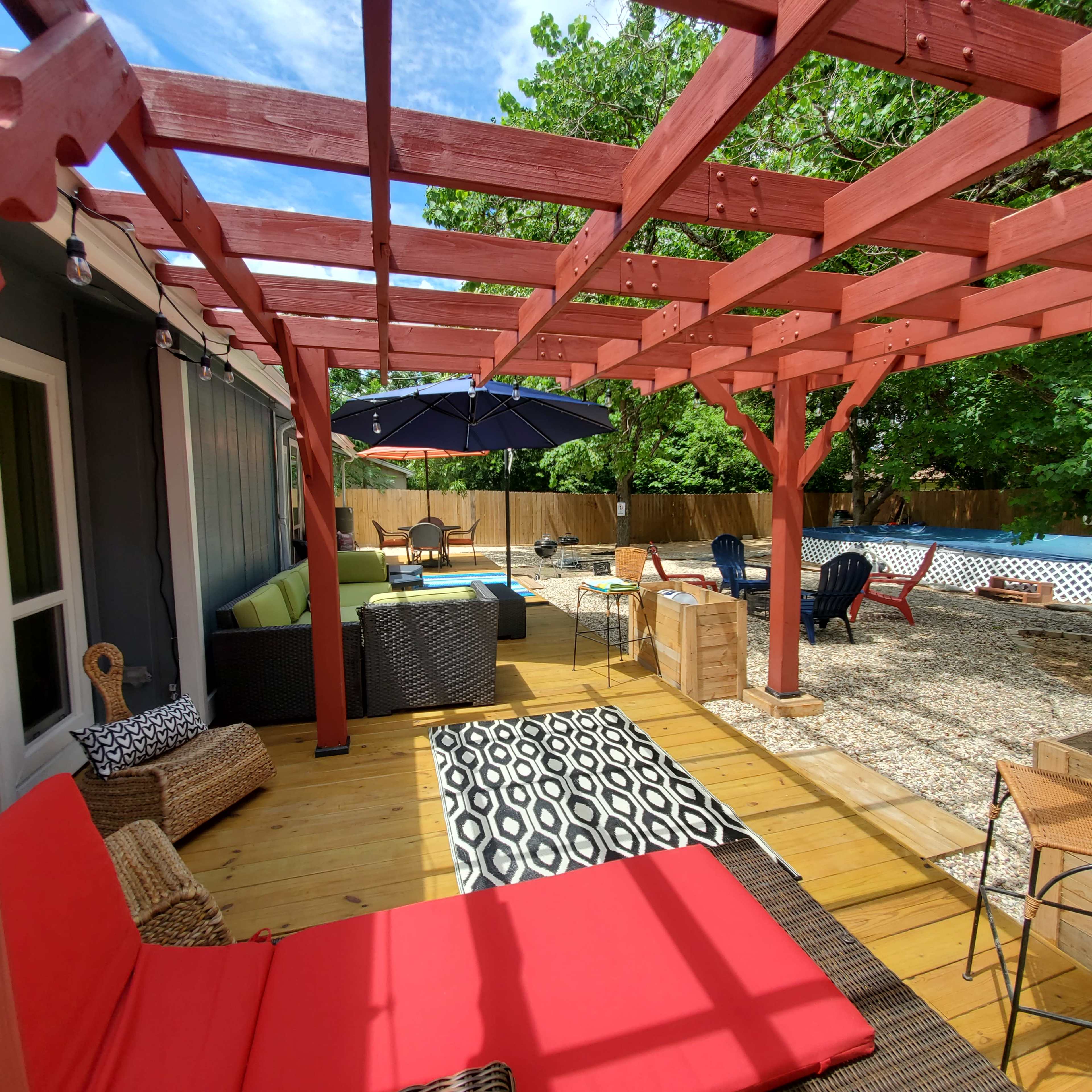 A spacious outdoor patio features a wooden pergola, colorful furniture, and a swimming pool in the background.