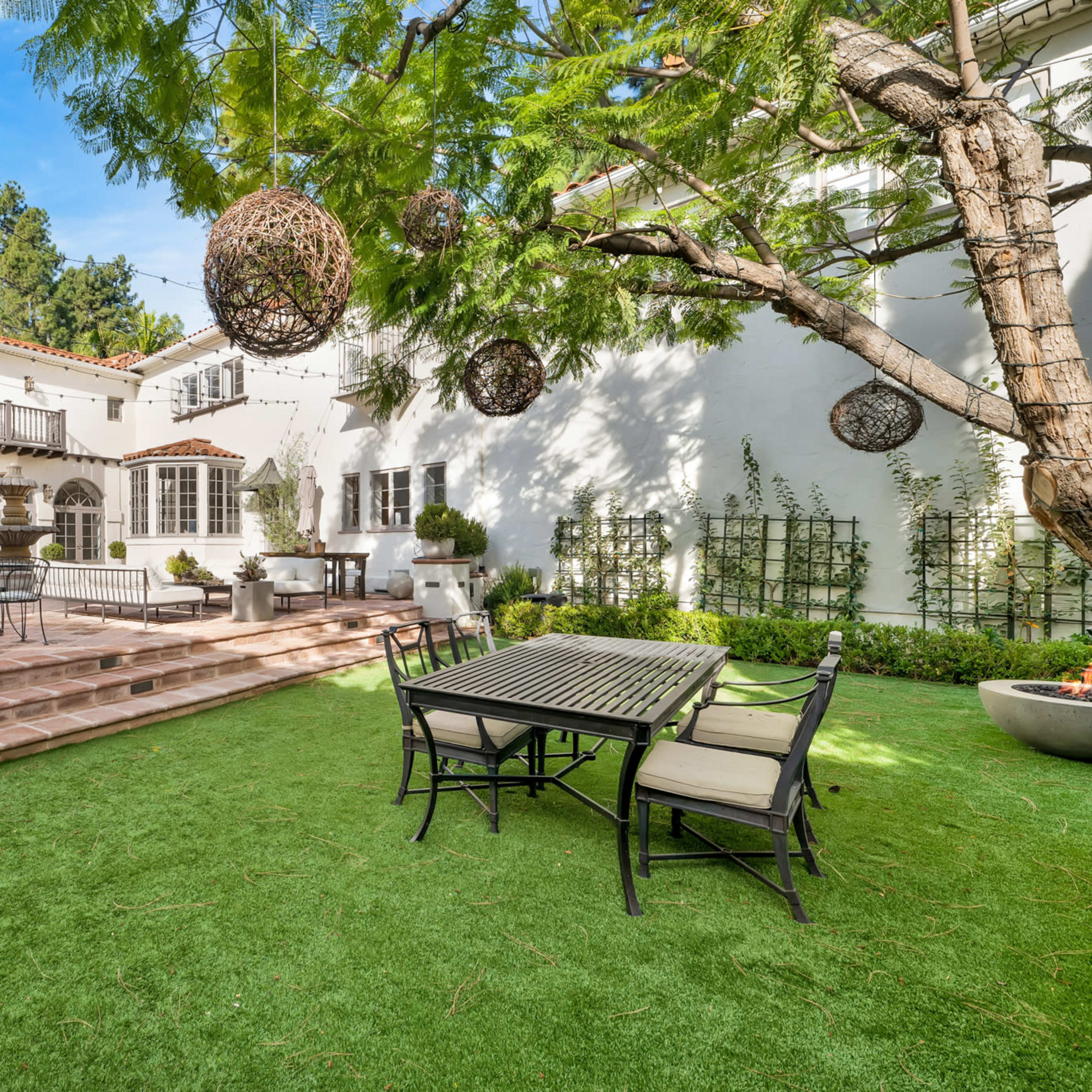 A landscaped outdoor patio area featuring a stone terrace, dining table, and a large tree with hanging decorations.