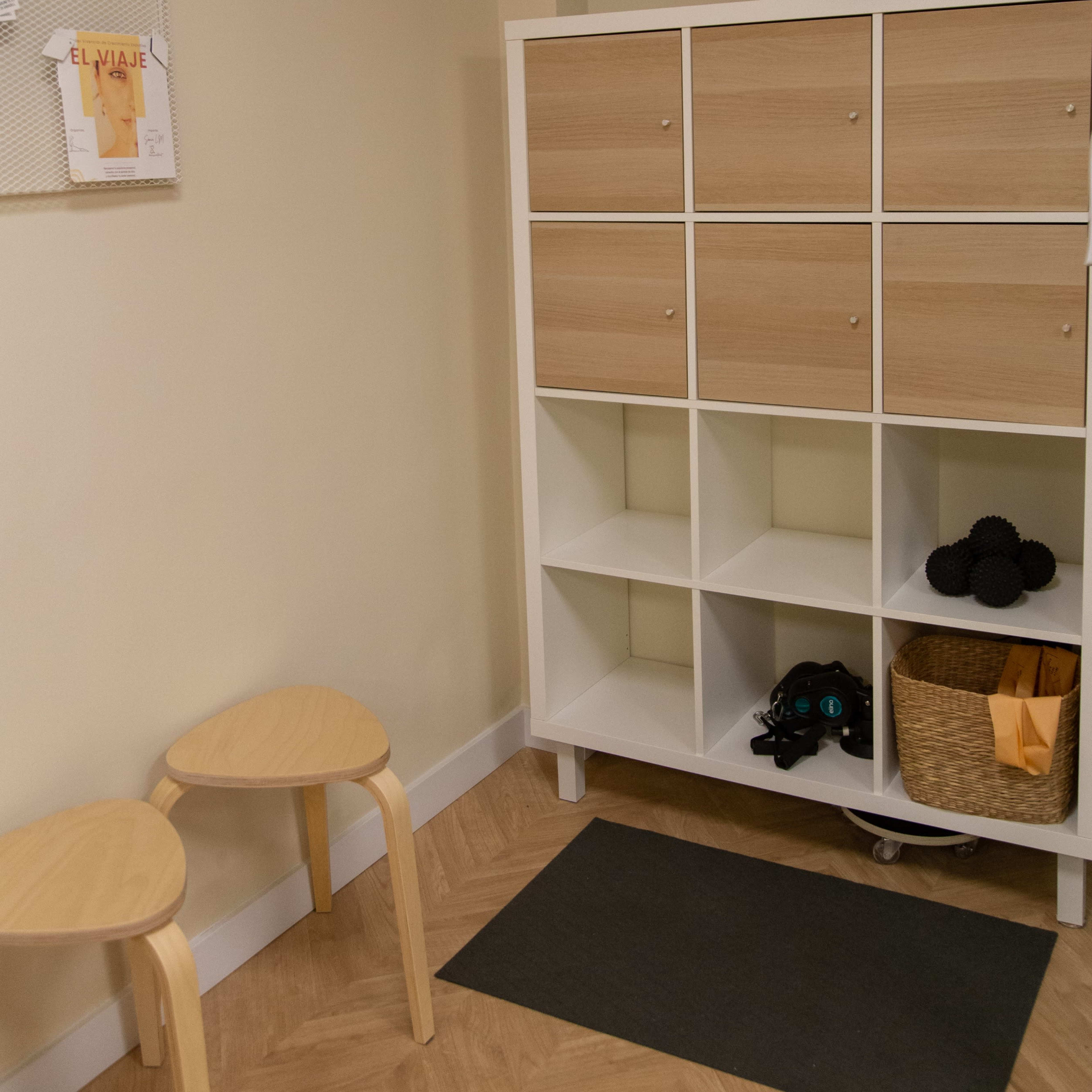 A small room with a white storage unit featuring cubbies and a basket, two wooden stools, and a wall with a notice board.