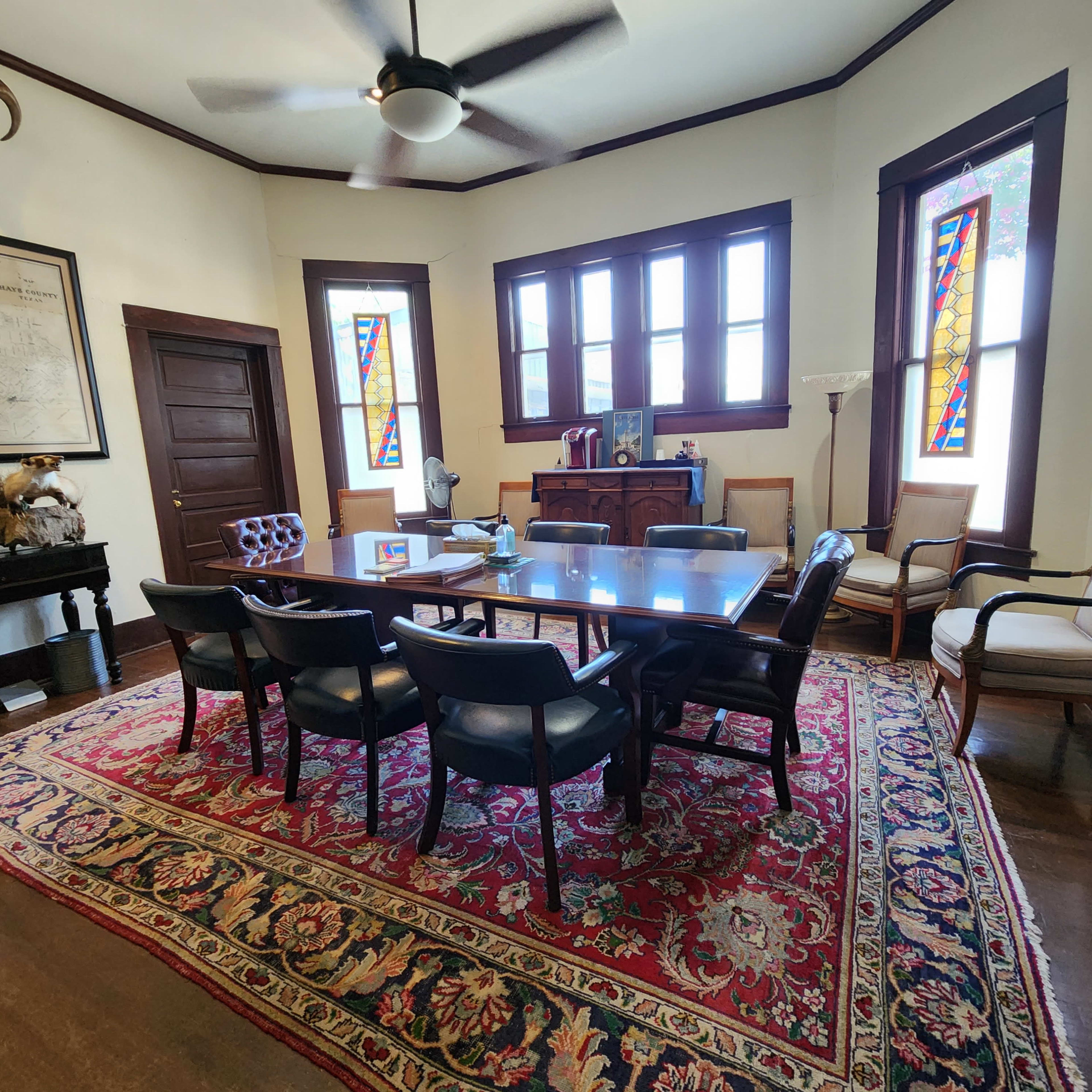A conference room features a large wooden table surrounded by black chairs, accented by a patterned rug, with stained glass windows and various pieces of furniture along the walls.