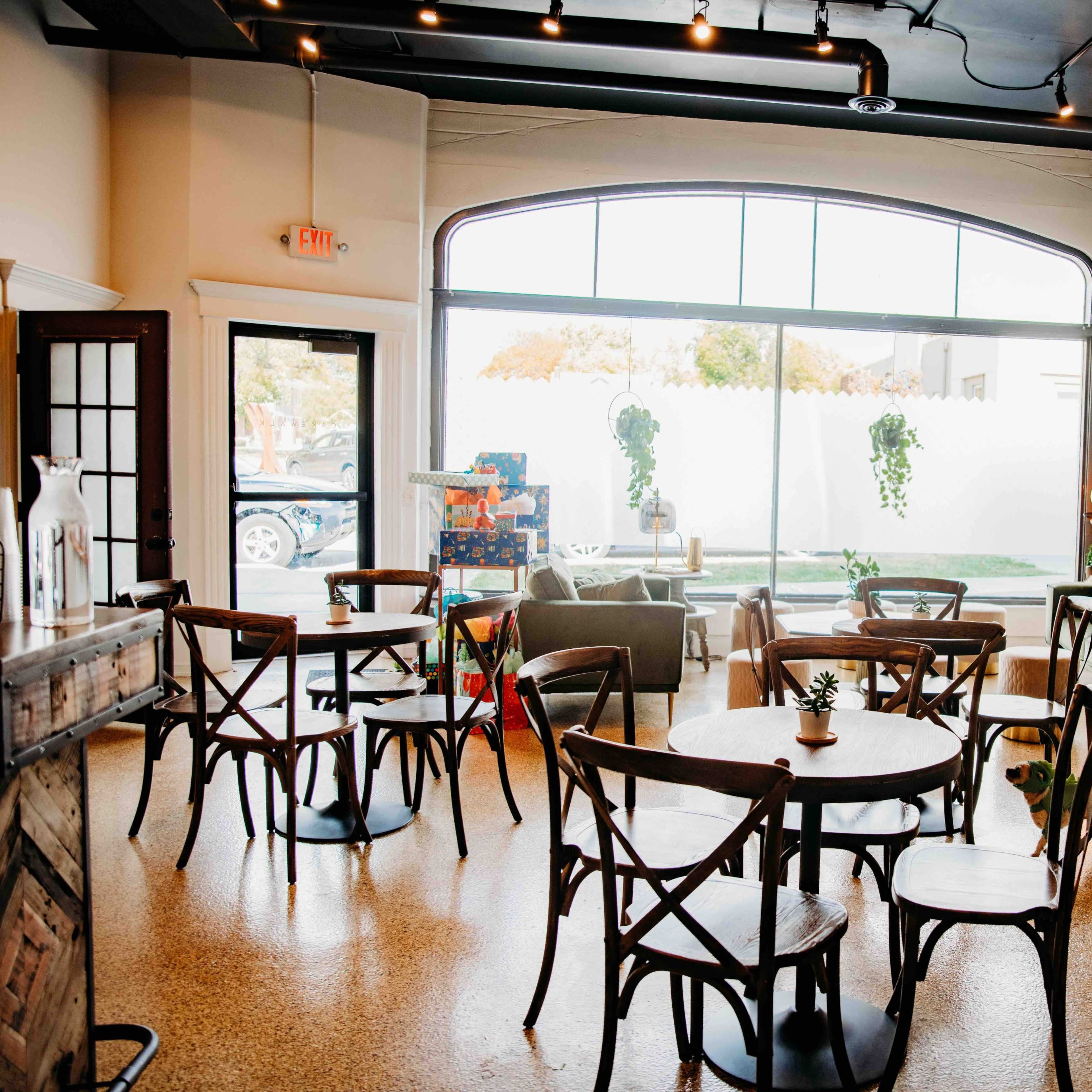 A cozy café interior with several wooden tables and chairs, a large window, and decorative plants.