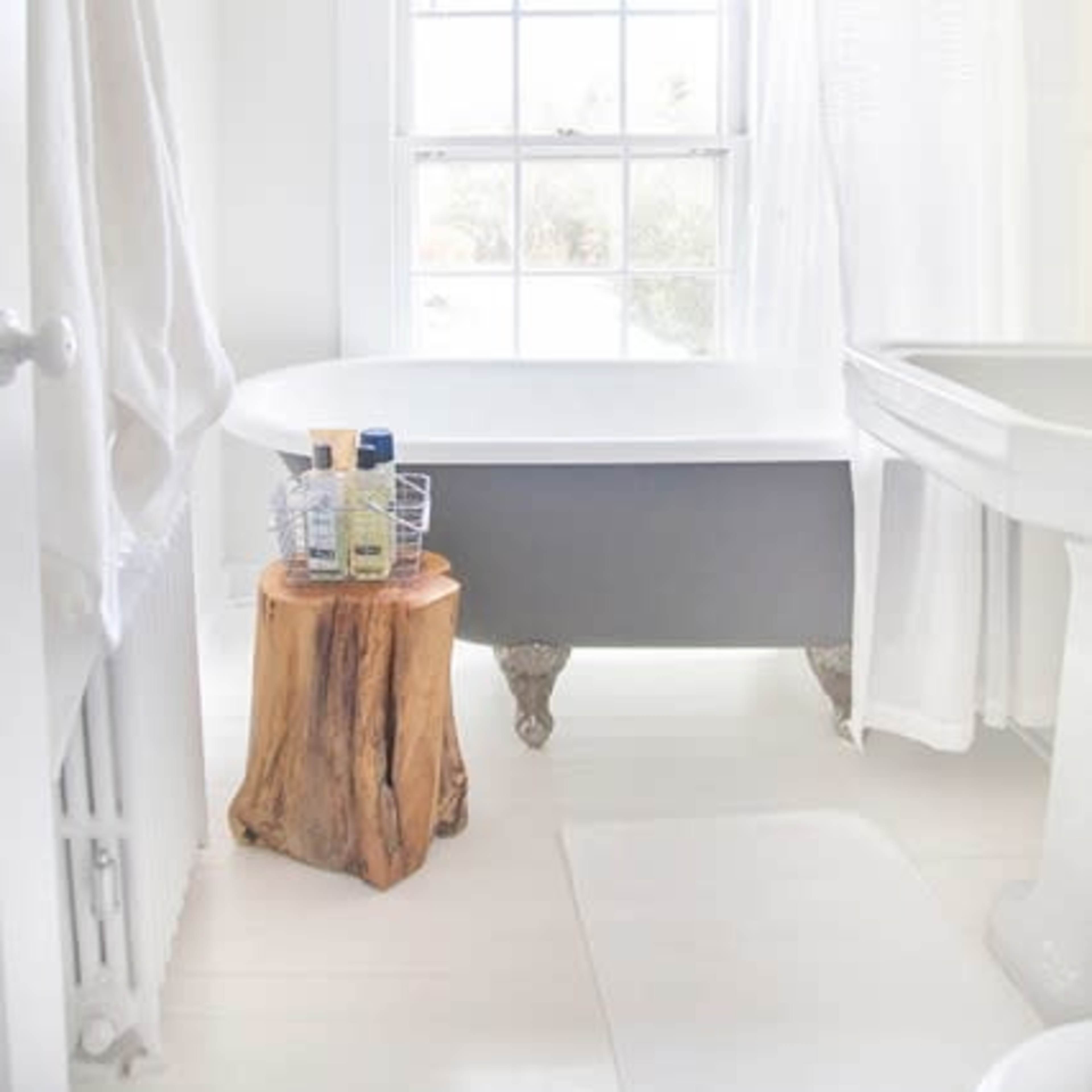 A bright and minimalistic bathroom featuring a freestanding bathtub, a wooden stool, and a window with sheer white curtains.