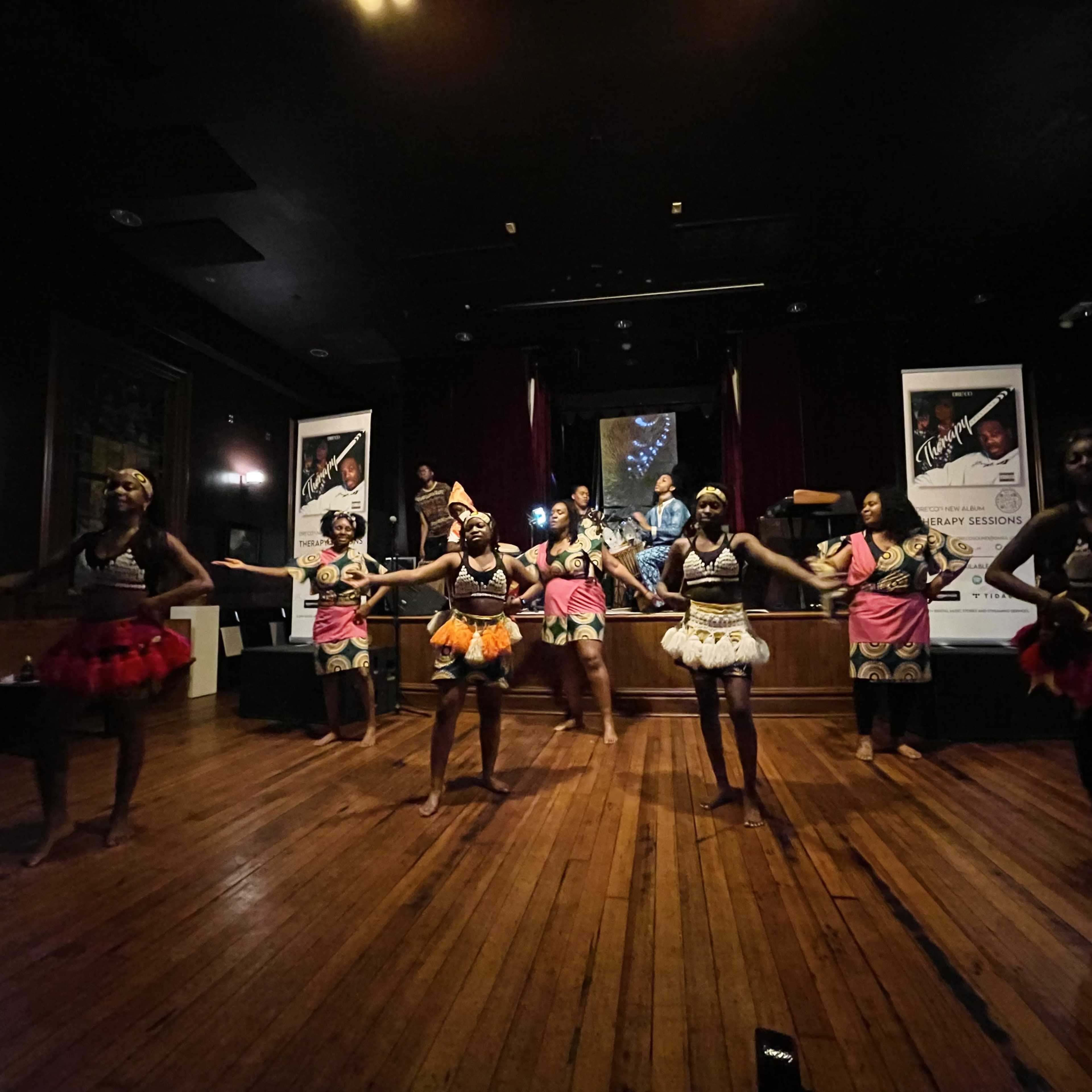 A group of dancers performs on stage wearing traditional attire in a dimly lit venue with posters in the background.