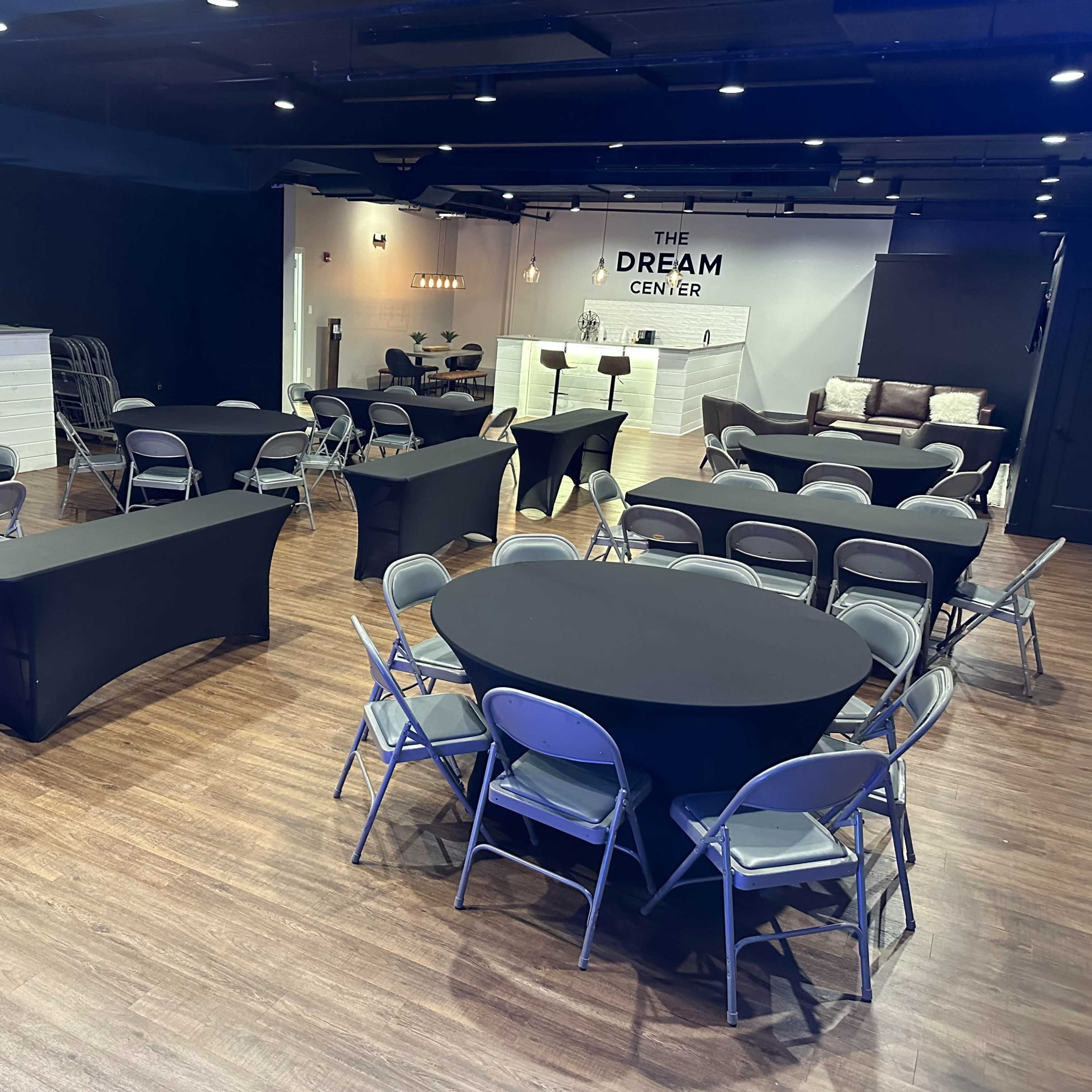 A spacious event room set with multiple round and rectangular tables covered in black tablecloths and accompanied by silver folding chairs.