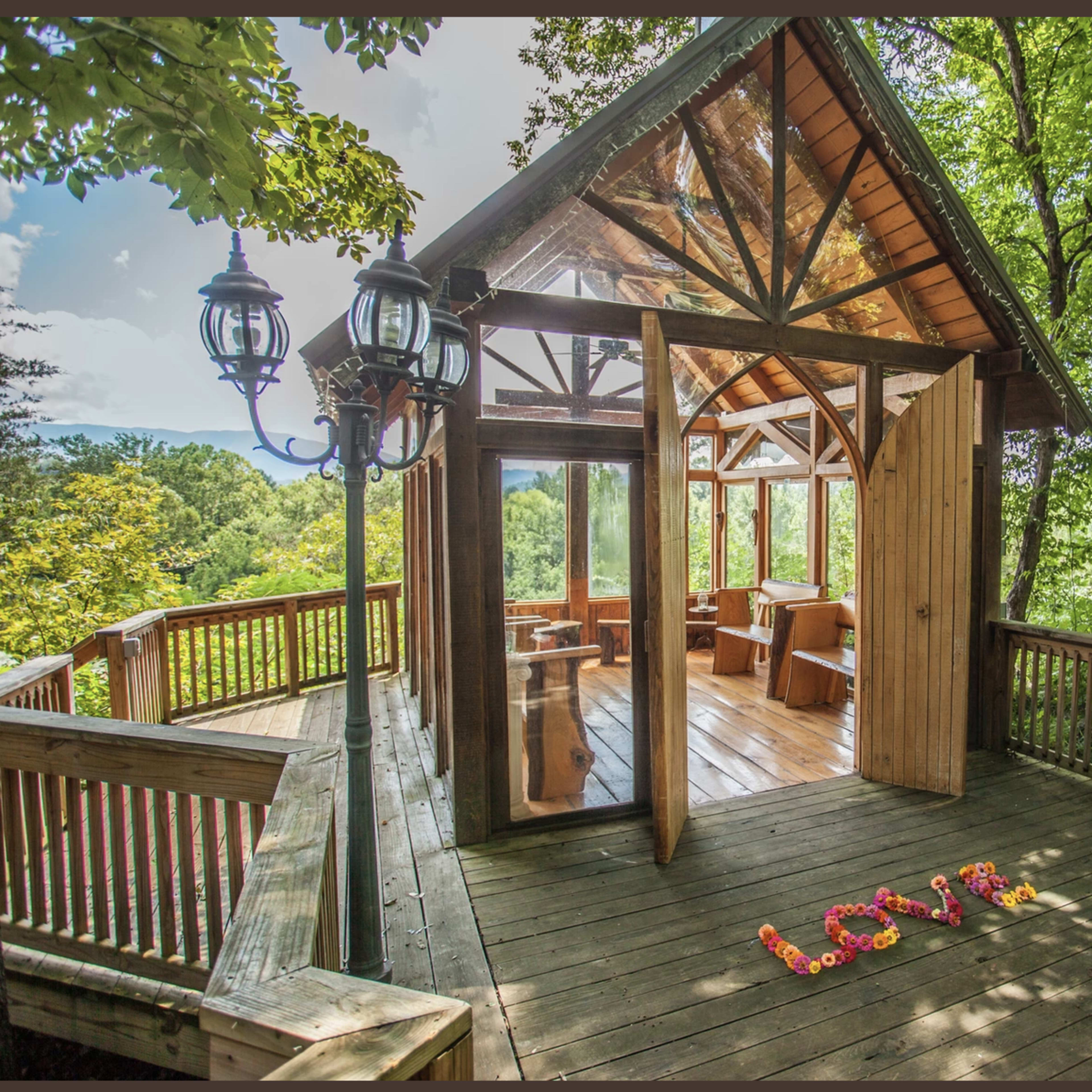 A wooden deck features a glass-paneled structure surrounded by trees, with the word "LOVE" spelled out in colored flowers on the floor.