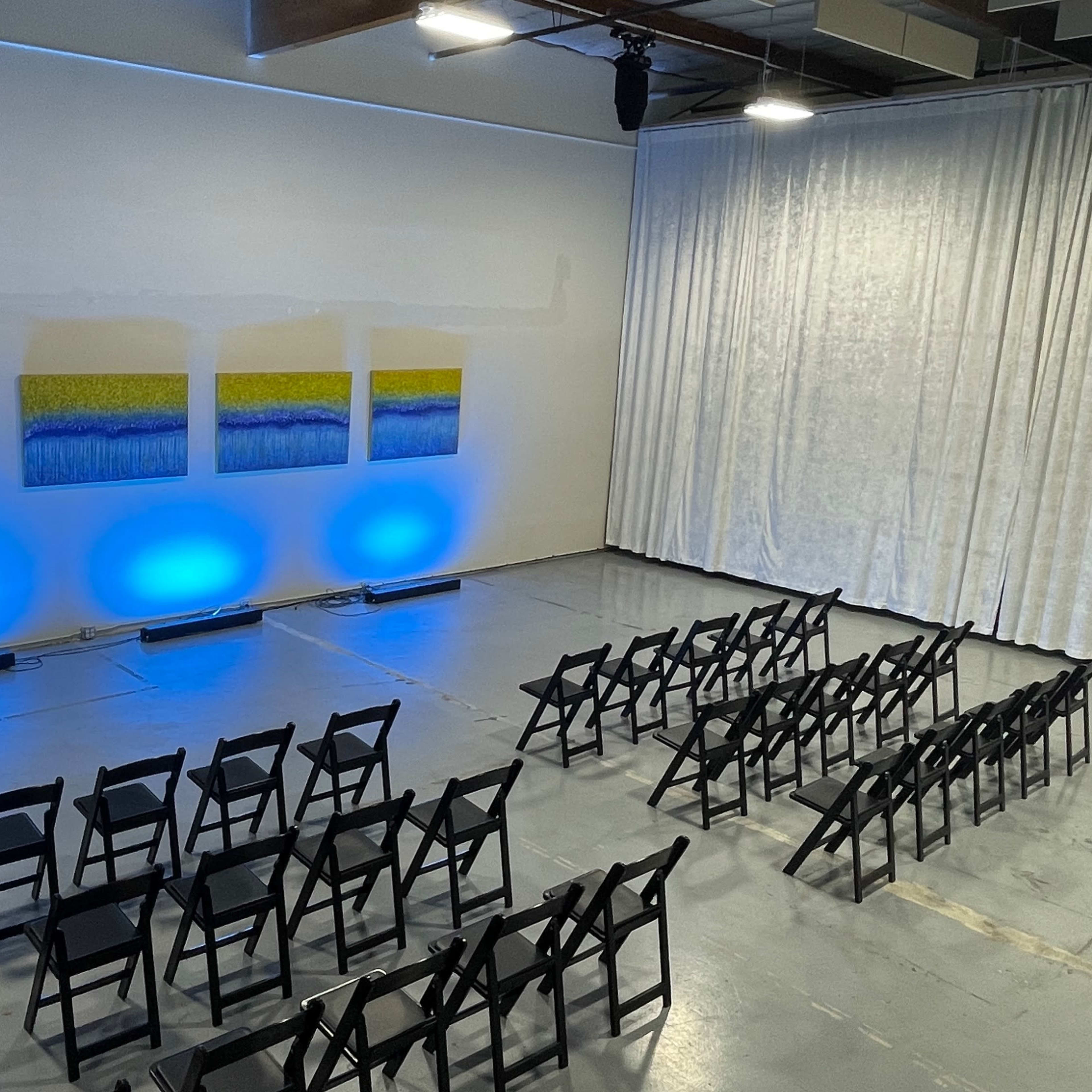 A spacious room features black chairs arranged in rows facing three large paintings illuminated by blue lights against a curtain backdrop.