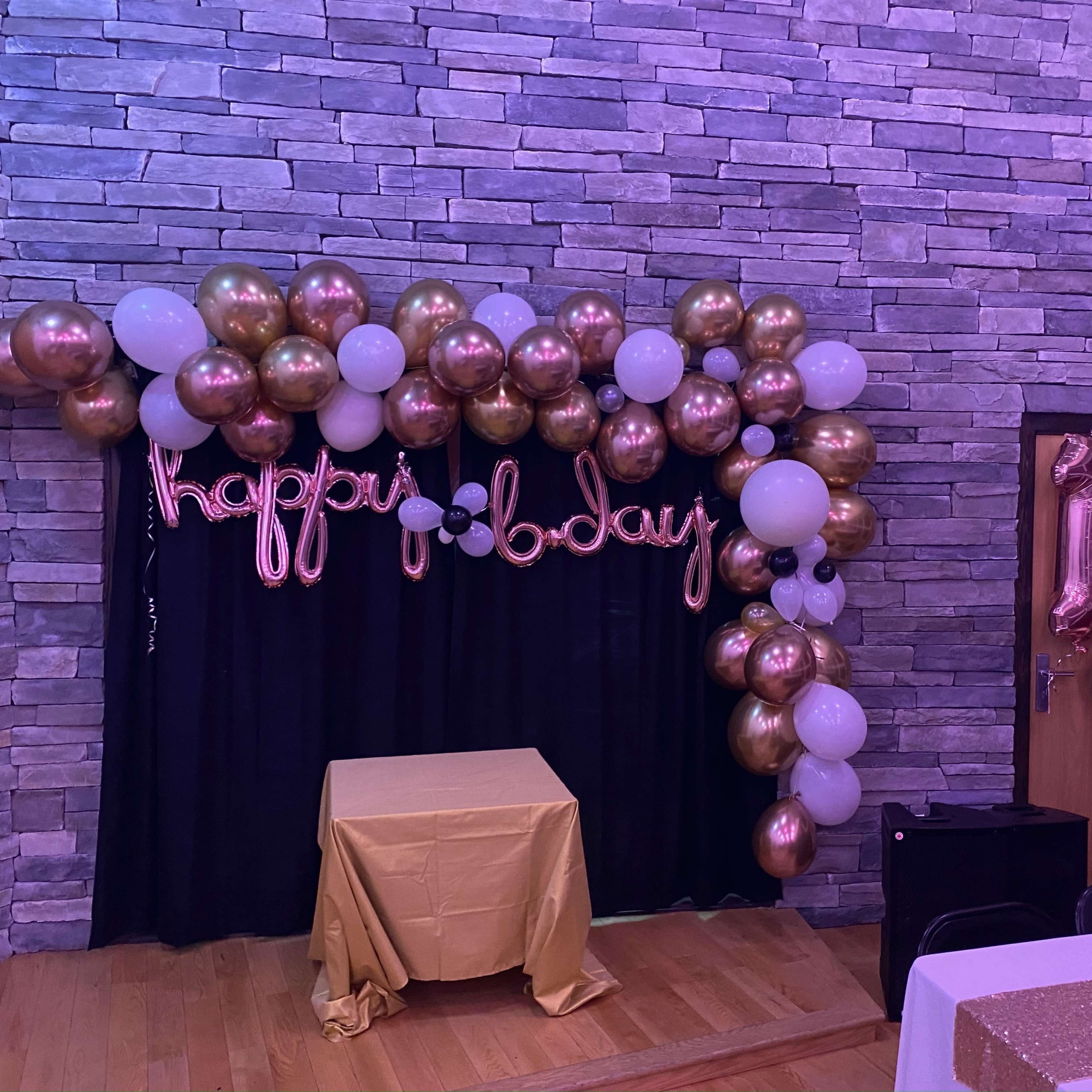 A decorated corner of a room featuring a backdrop with balloons, a "happy 18th birthday" banner, and a table draped with a gold tablecloth.