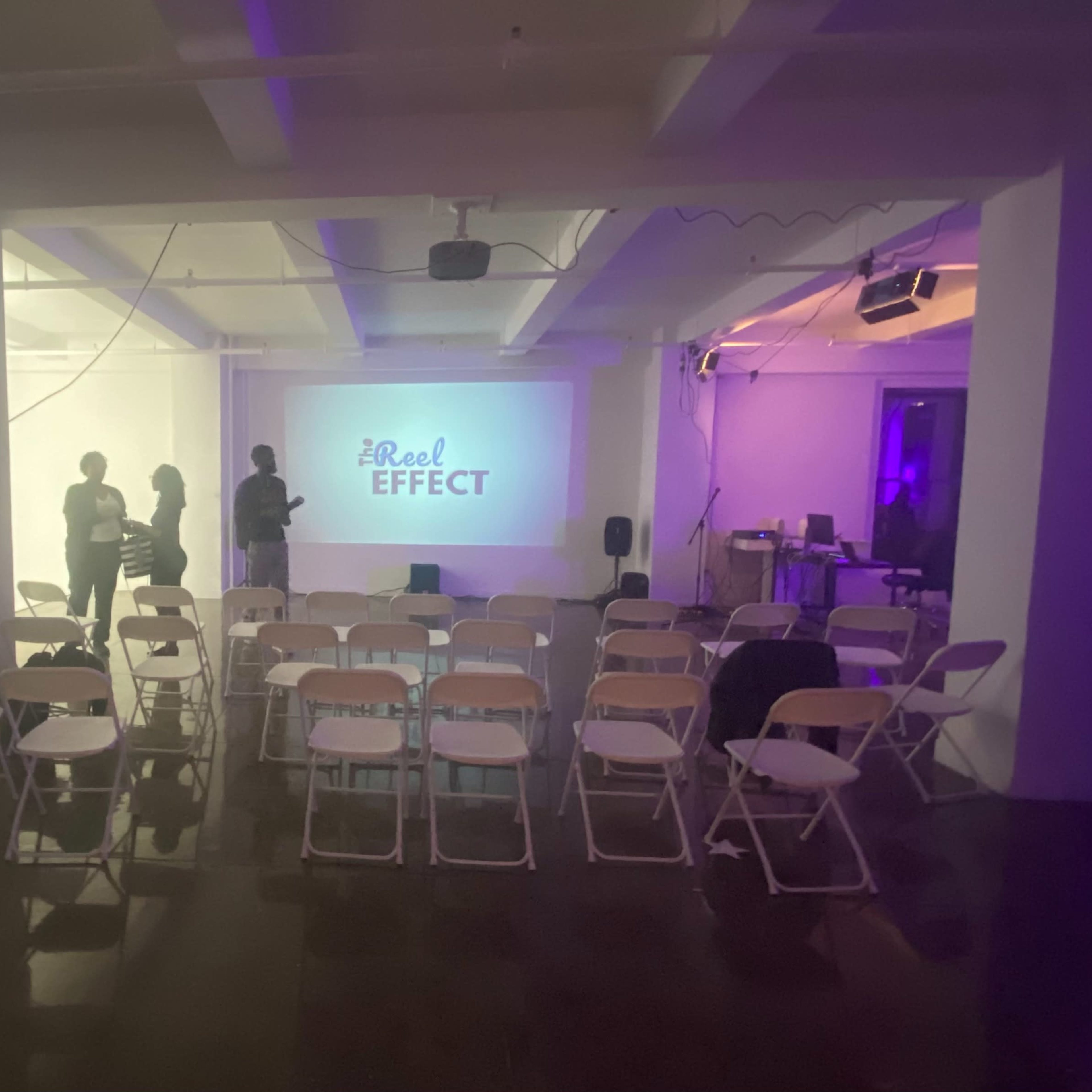 A modern event space is set up with rows of chairs facing a screen displaying "The Reel Effect," while shadows of people are seen preparing for the event.