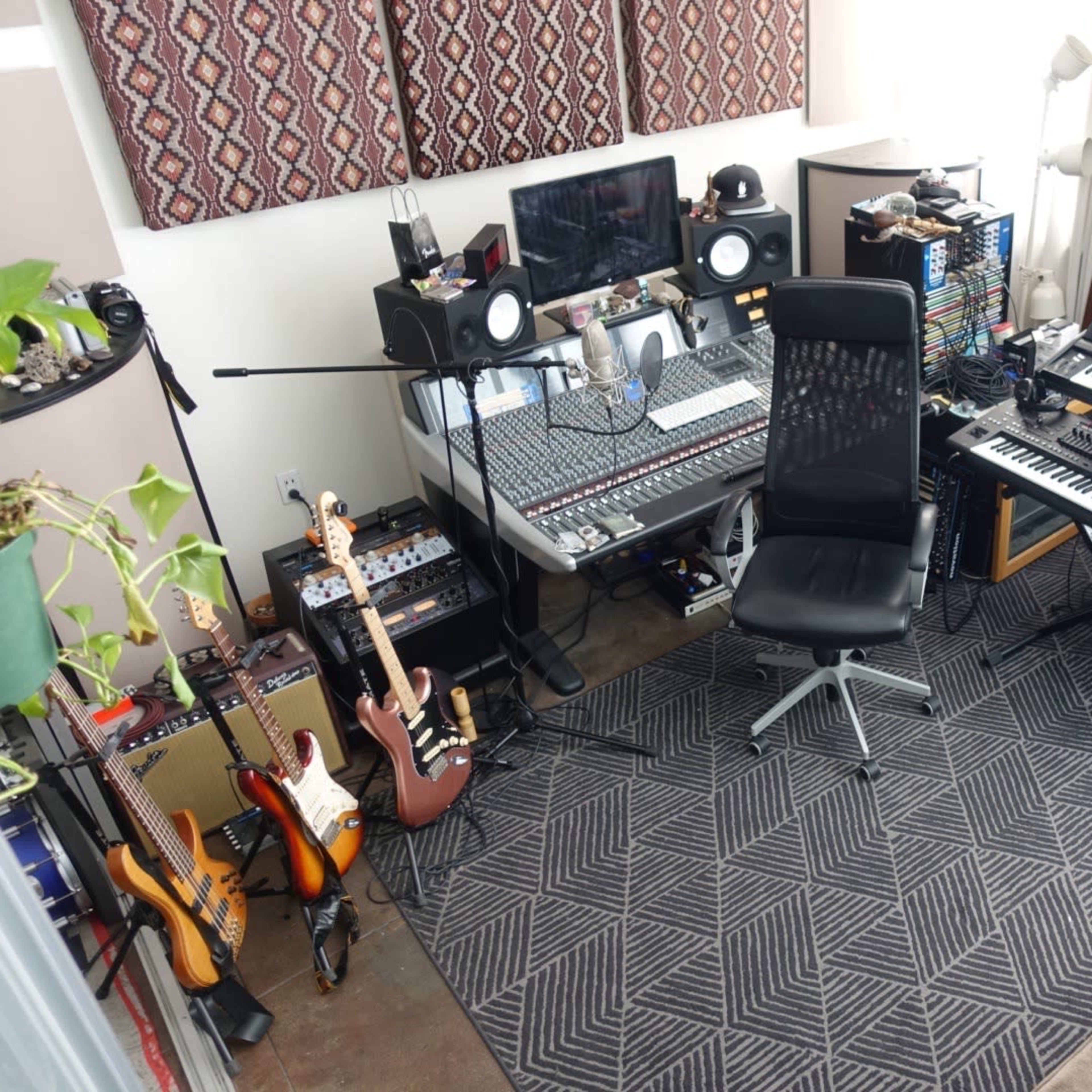 A music production room with a sound mixing console, keyboard, various musical instruments, and a workstation chair arranged around a cozy seating area.