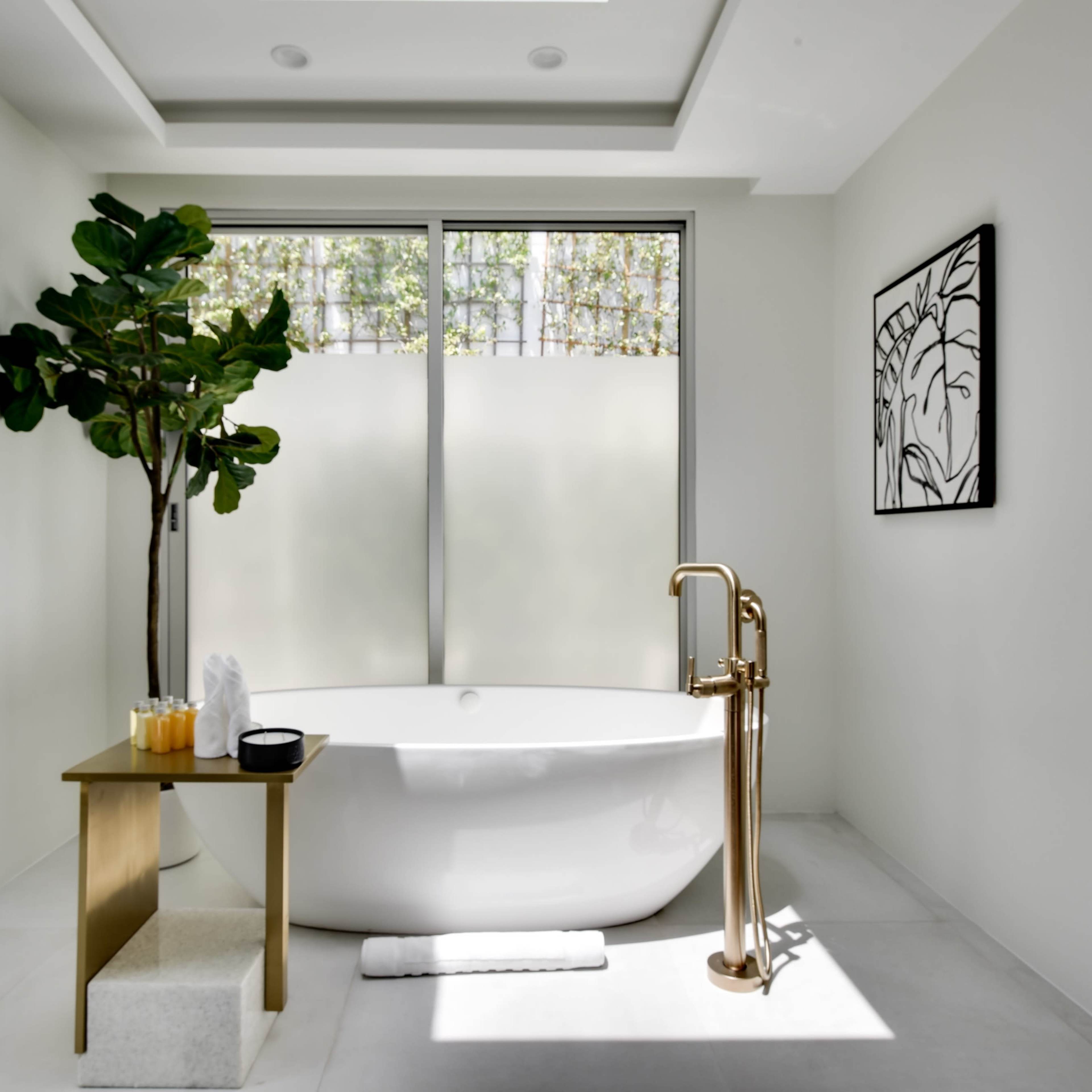A modern bathroom features a freestanding bathtub, a floor lamp, and a window with frosted glass, along with a potted plant and a framed artwork on the wall.