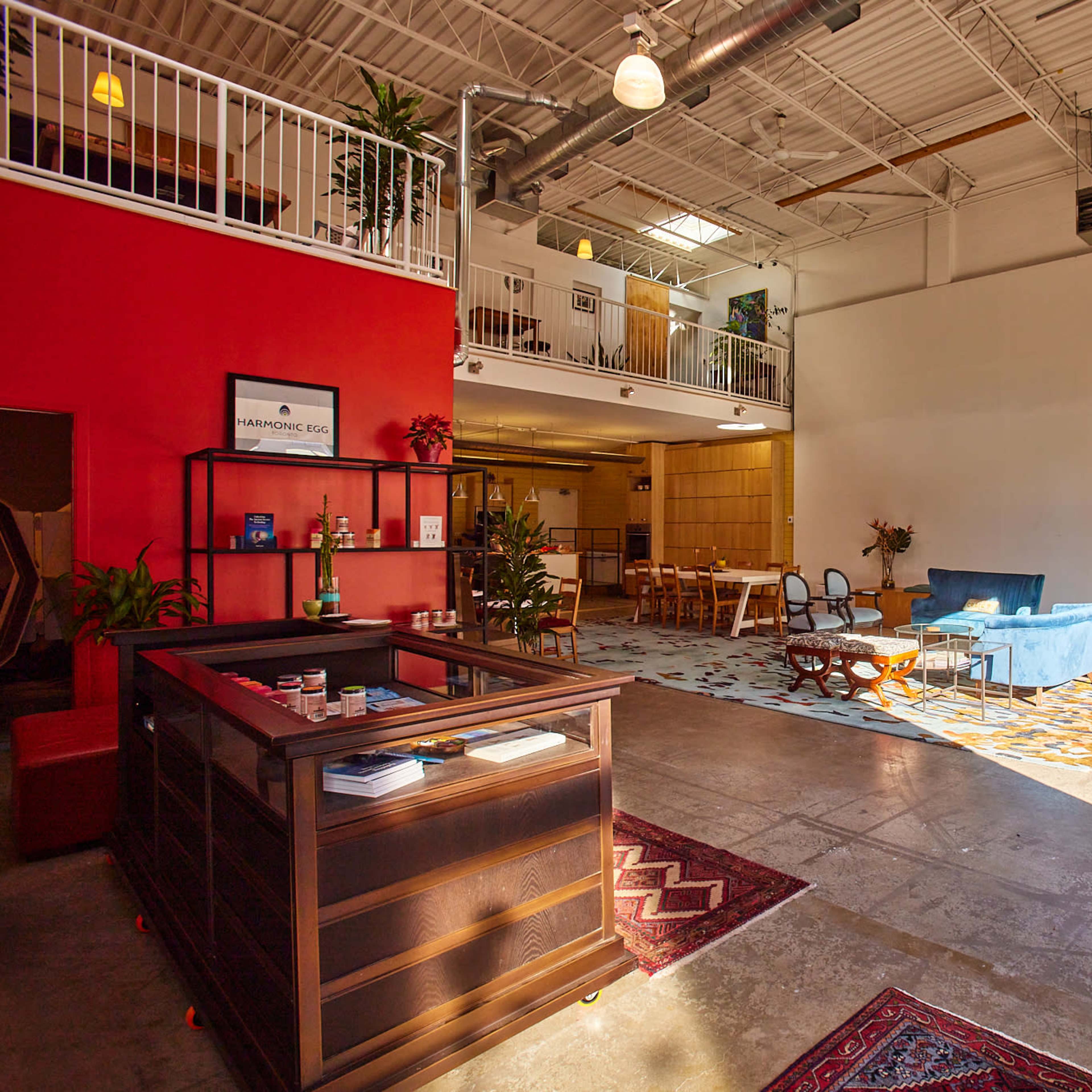 An open workspace with high ceilings, featuring a reception area, a seating area with blue furniture, and a dining space in the background.