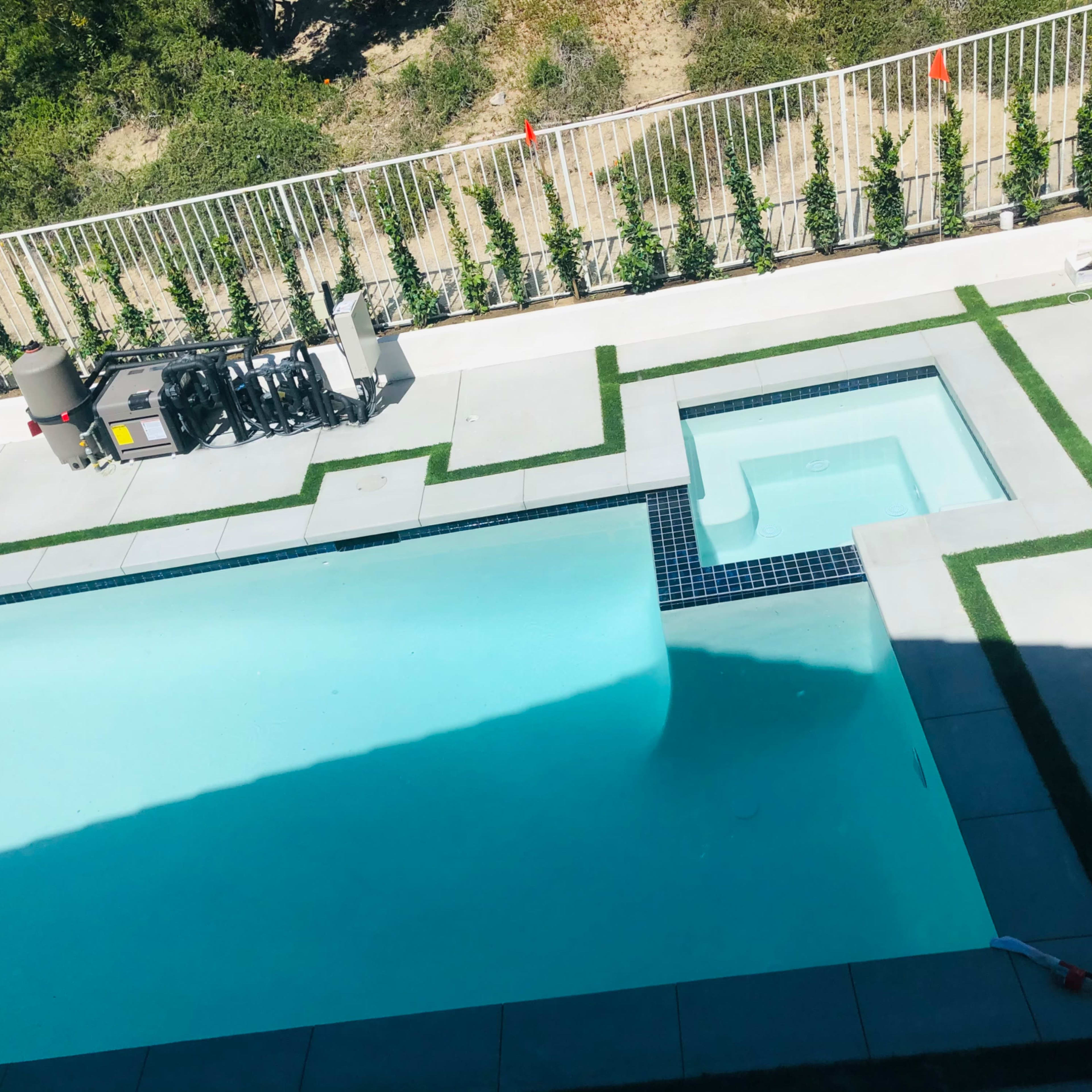 A modern swimming pool with a connected hot tub, surrounded by a concrete patio and a green lawn border.