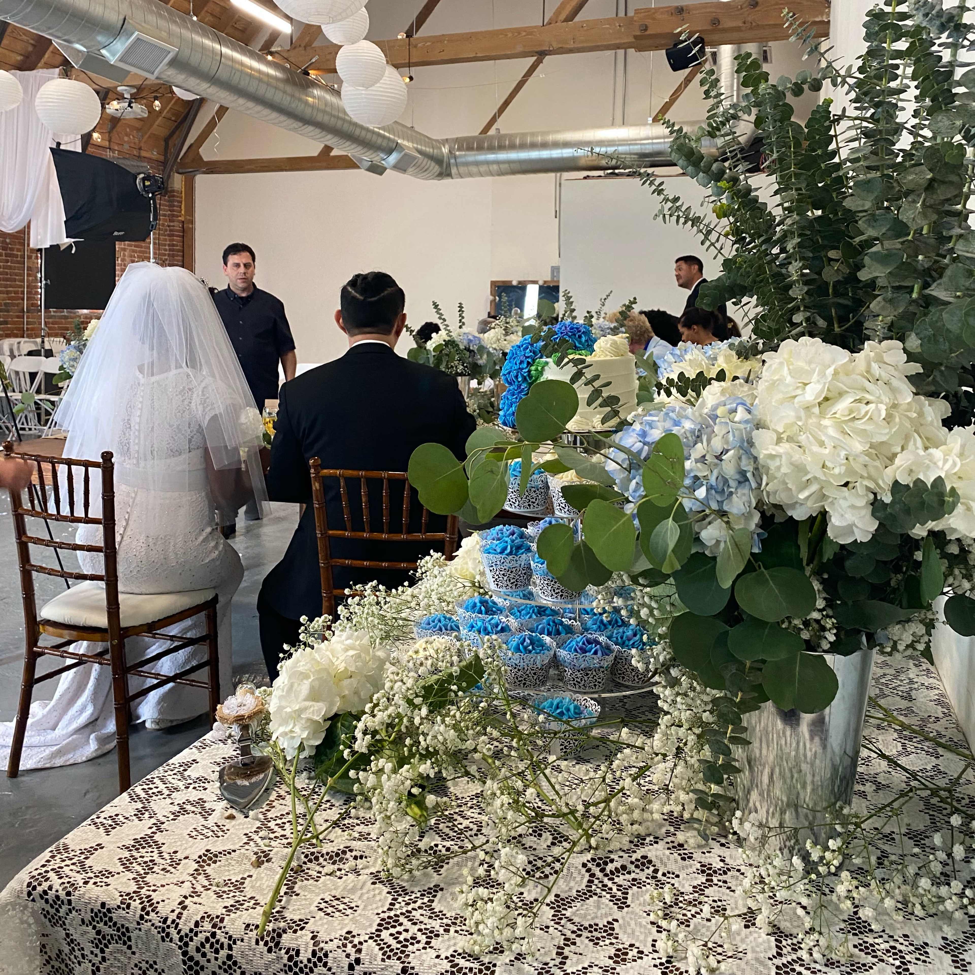 A bride in a white dress sits at a table decorated with flowers and greenery while a person stands, addressing the small gathering in an indoor venue.