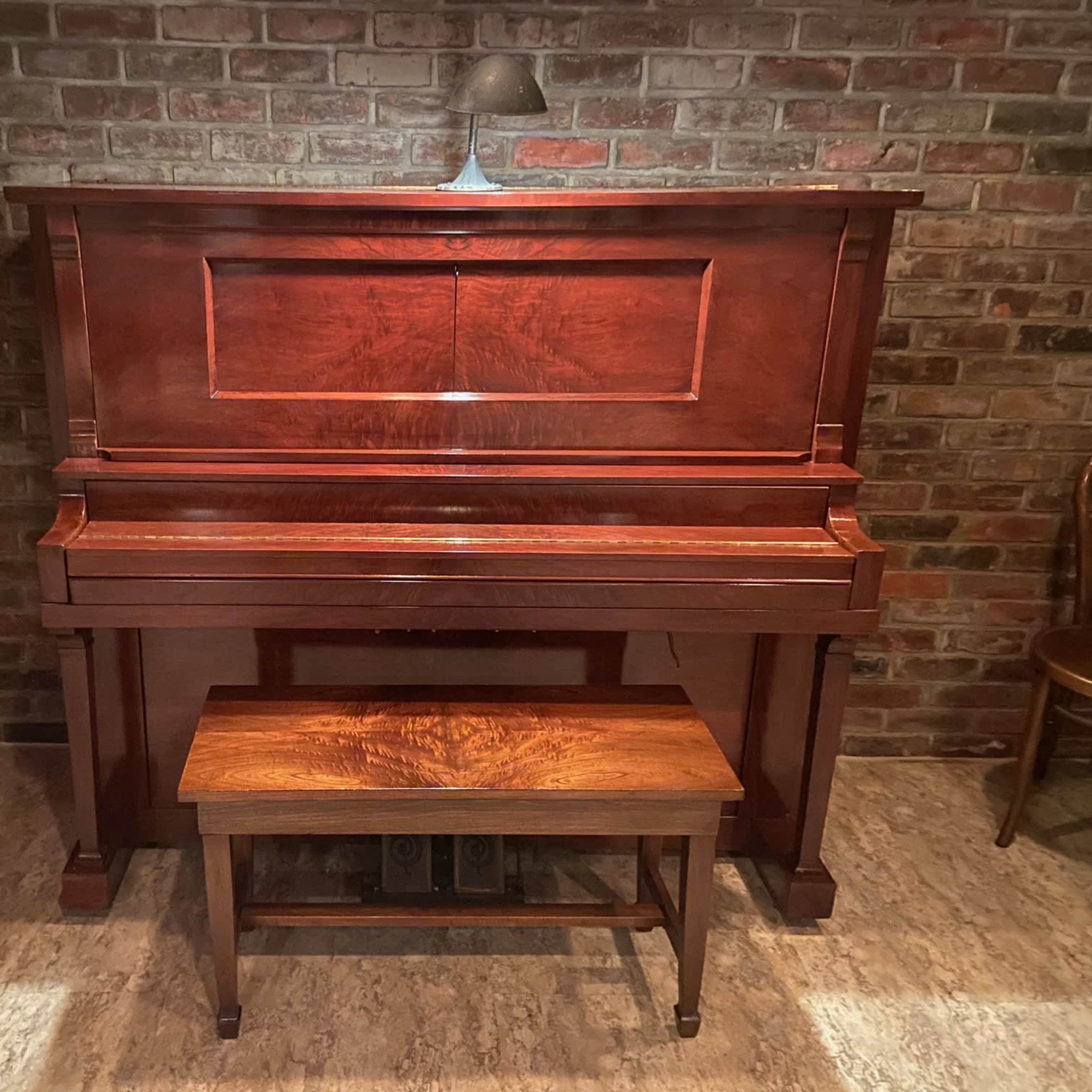 A wooden piano with a matching bench is positioned against a brick wall, illuminated by a small lamp.