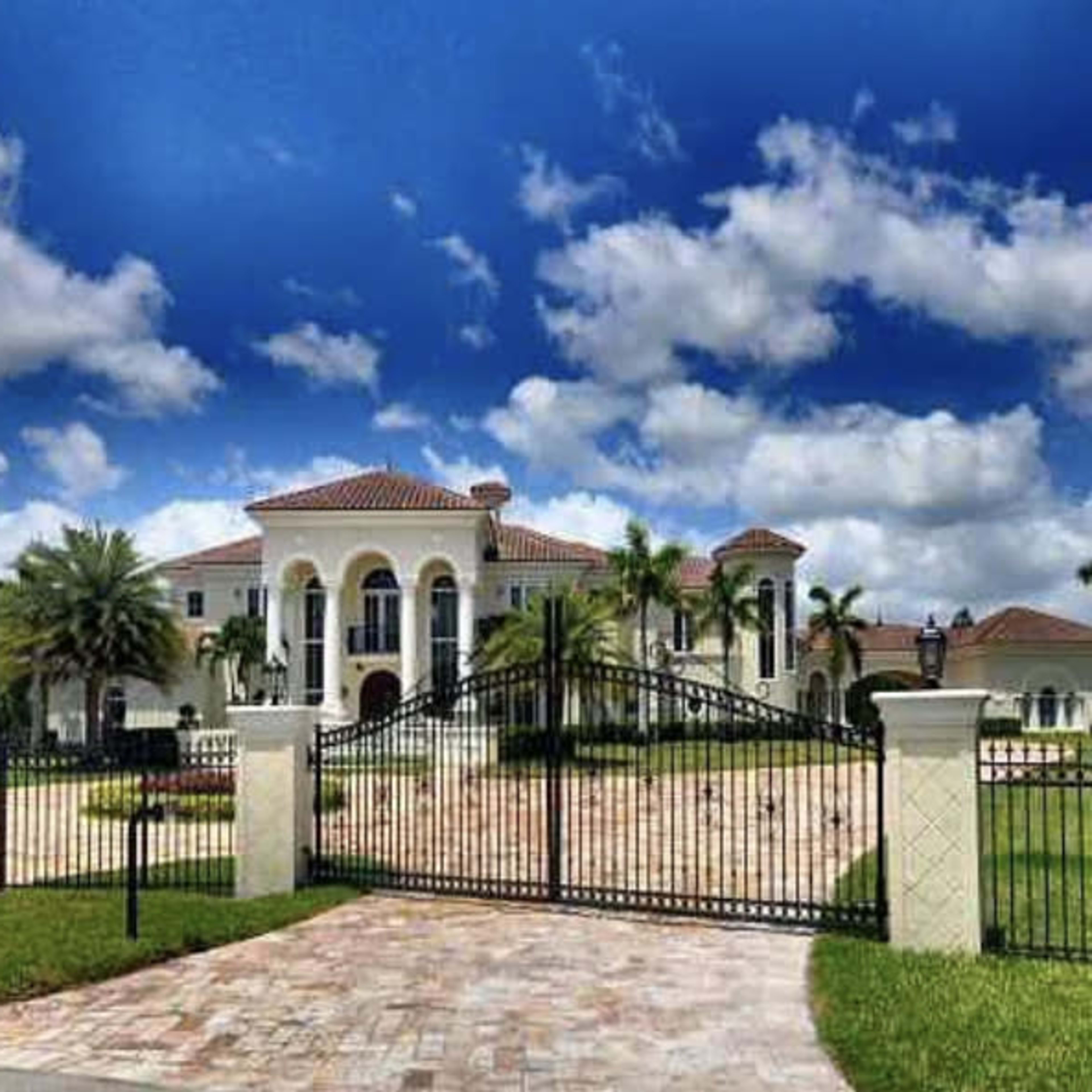 A large, two-story mansion with a red-tiled roof and palm trees is enclosed by a black wrought-iron gate, set against a backdrop of blue sky and scattered clouds.