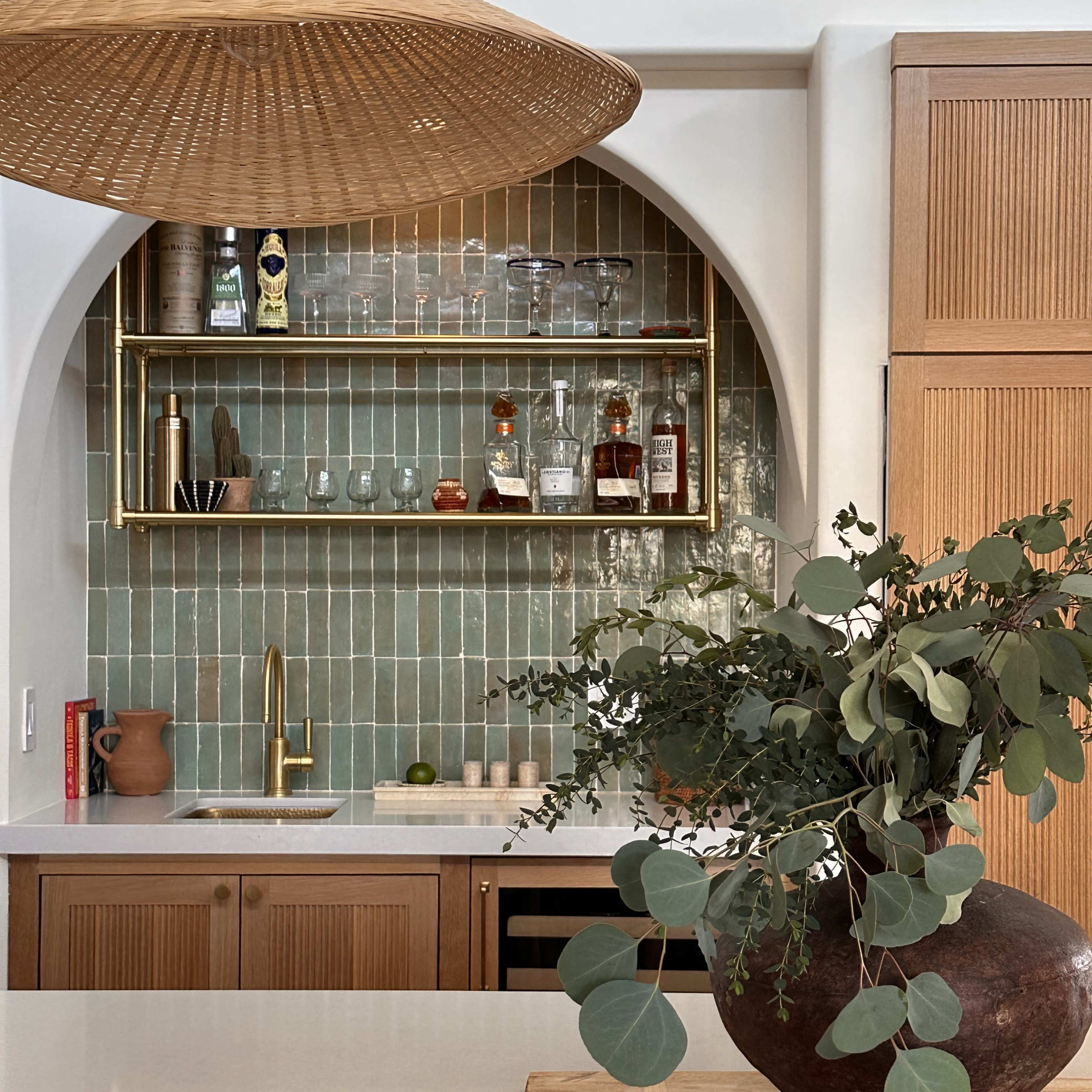 A modern kitchen features a large pendant light above a countertop with a vase of greenery, alongside shelves displaying glassware and bottles against a green tiled wall.