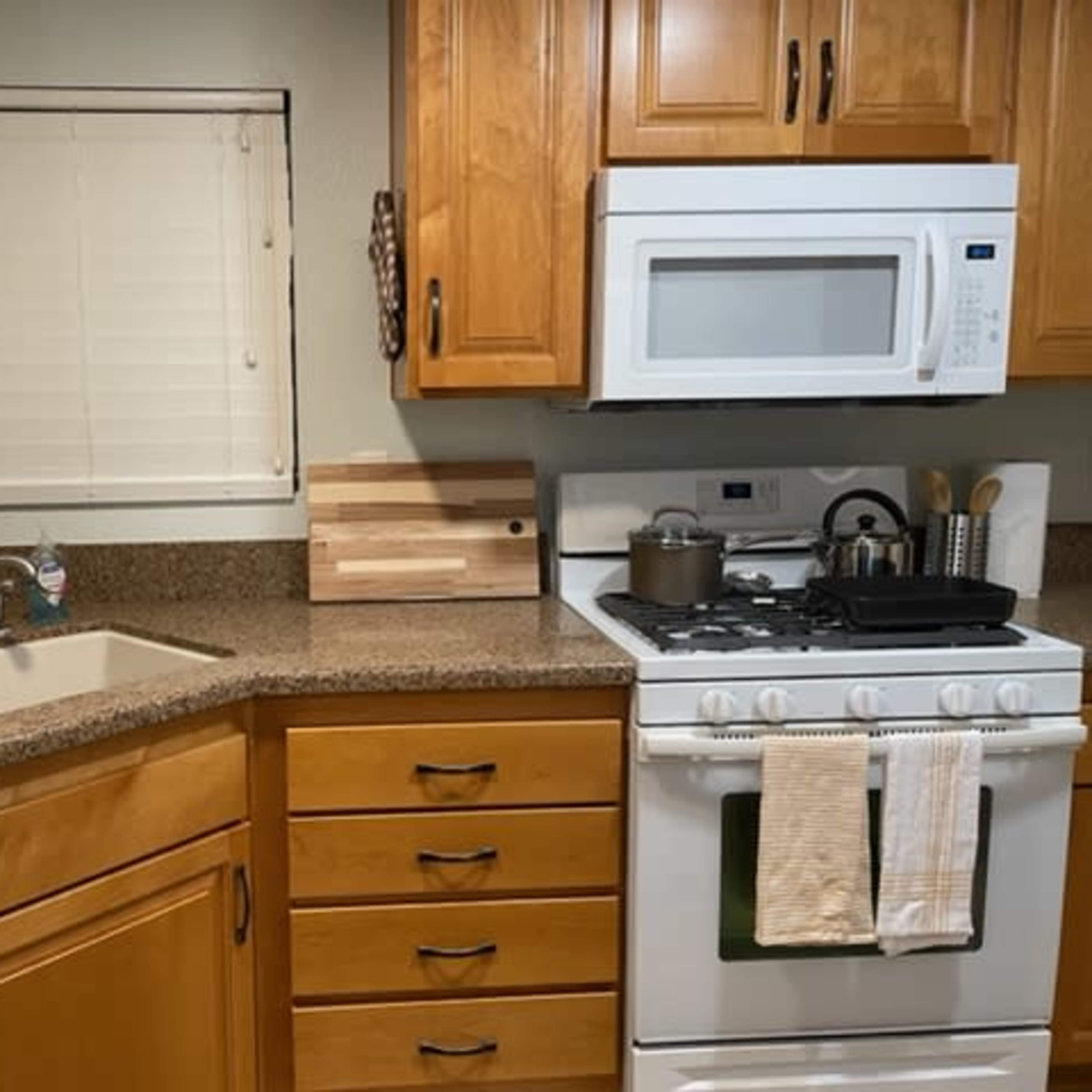 A kitchen with wooden cabinets, a double sink, a stove with a pot, and jars on the counter beside a window.