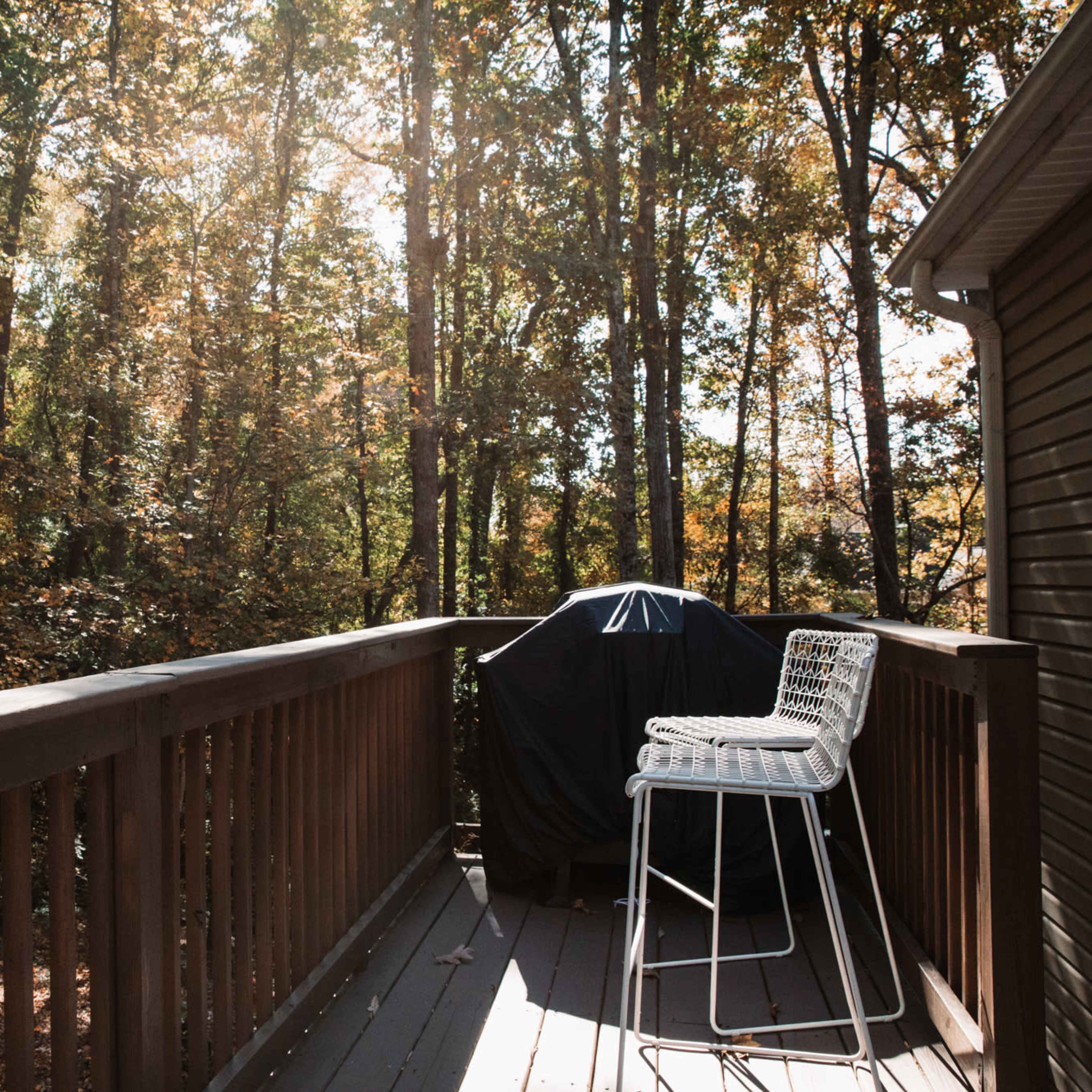 A wooden deck overlooks a wooded area with trees displaying autumn foliage, featuring a covered grill and a white bar stool.