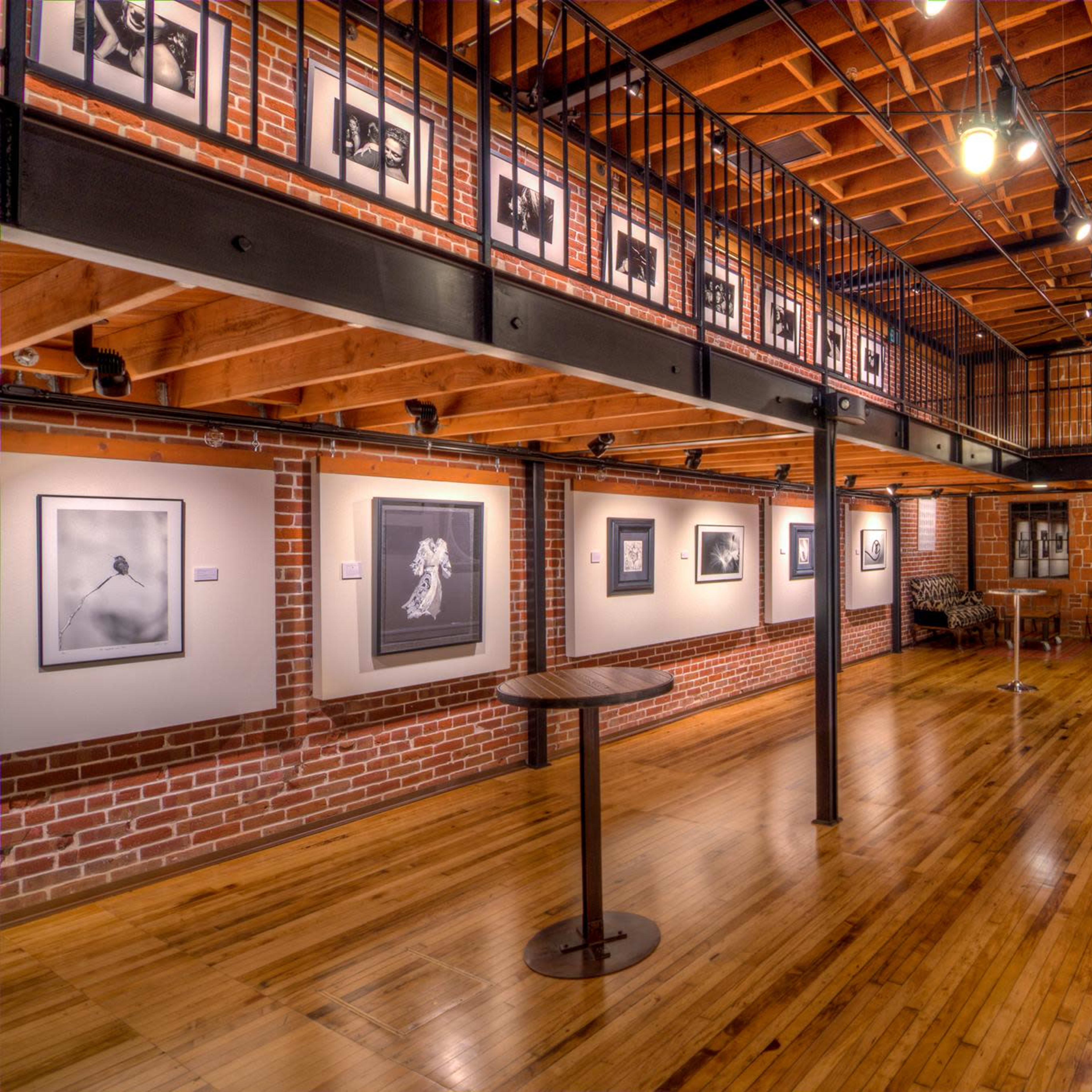 A contemporary art gallery with black-and-white photographs displayed on brick walls, wooden flooring, and a balcony overlooking the main exhibition area.