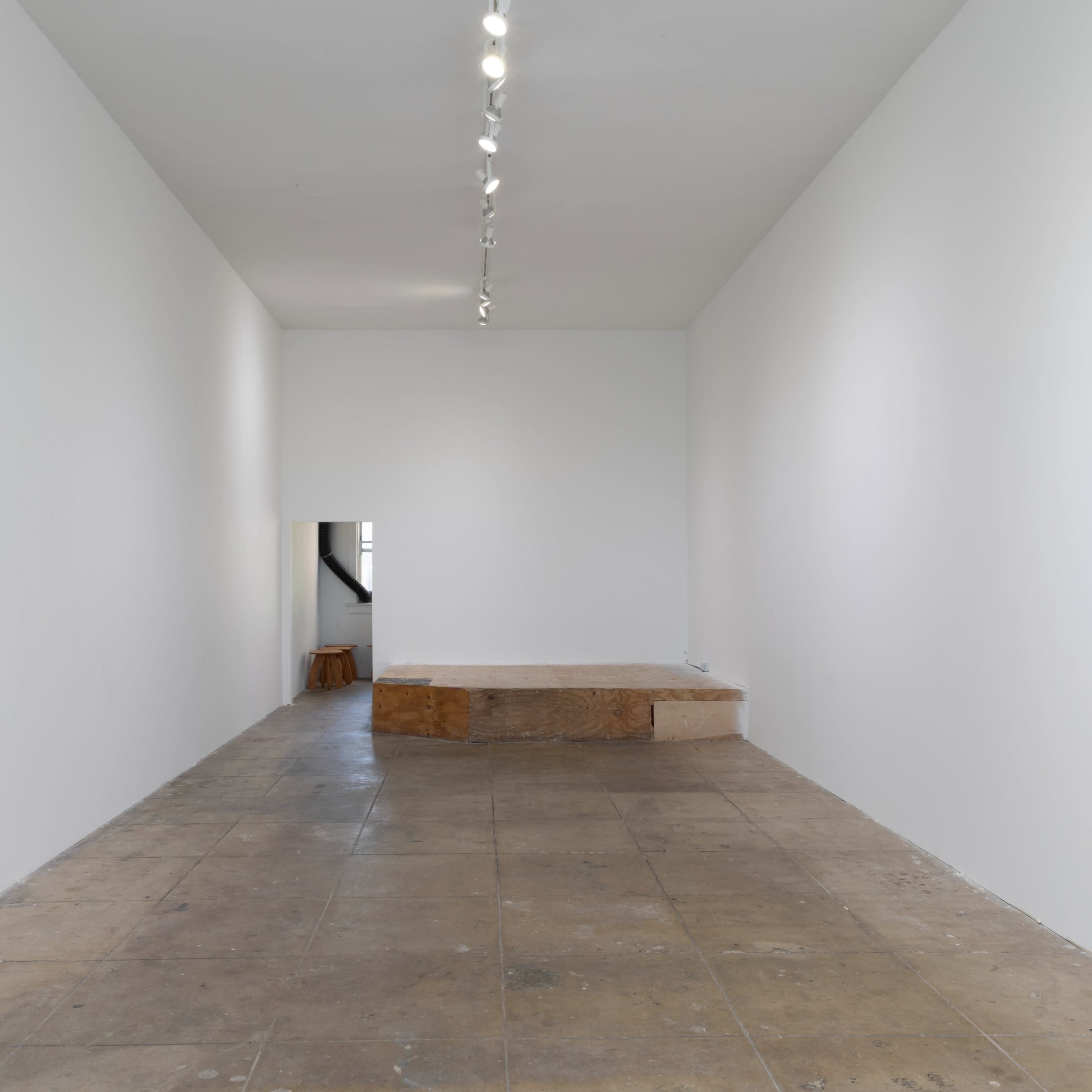 A minimalist art gallery featuring a stark white space with a wooden platform positioned against one wall.
