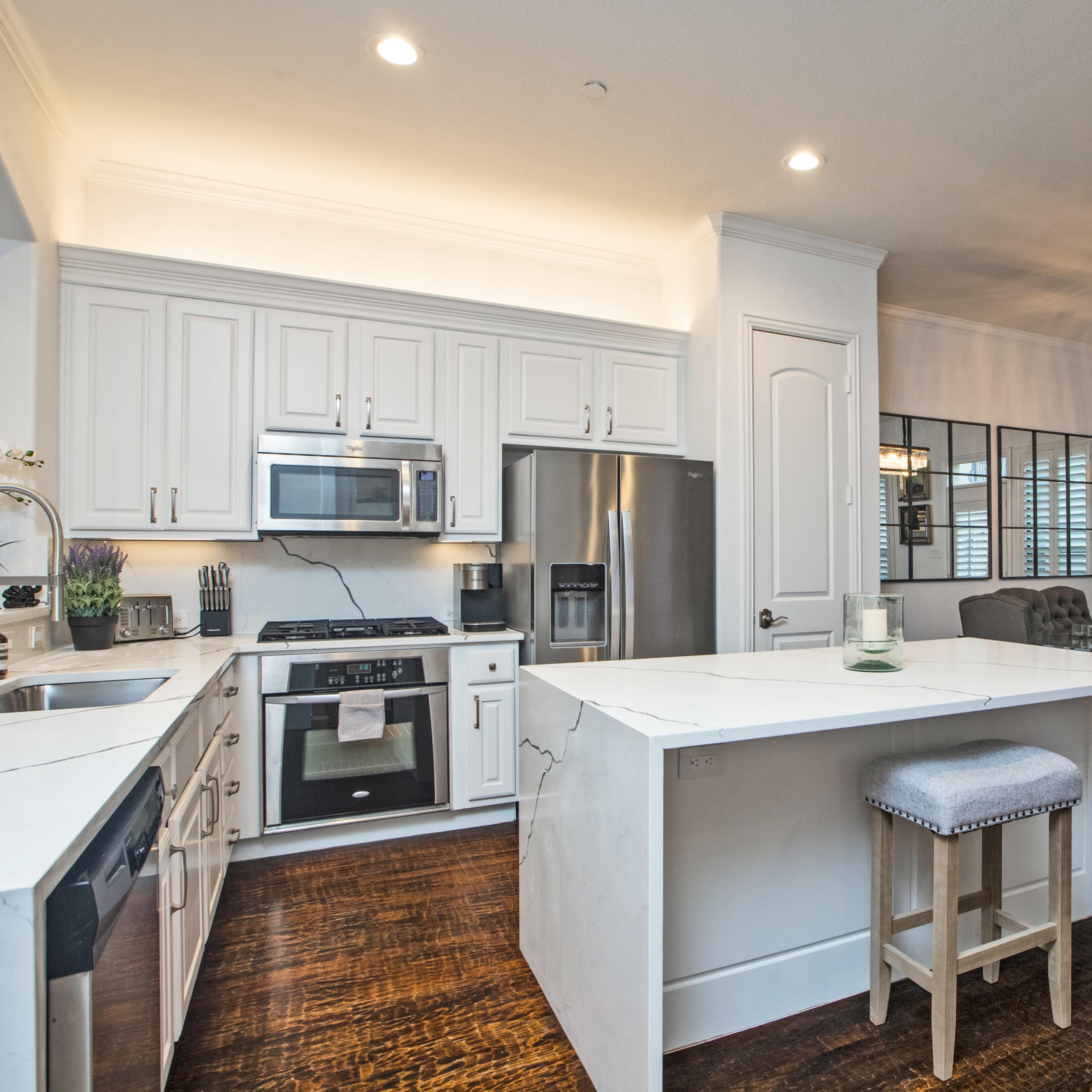 A modern kitchen with white cabinetry, stainless steel appliances, and an island with a seating area, adjacent to a dining area with a table and chairs.
