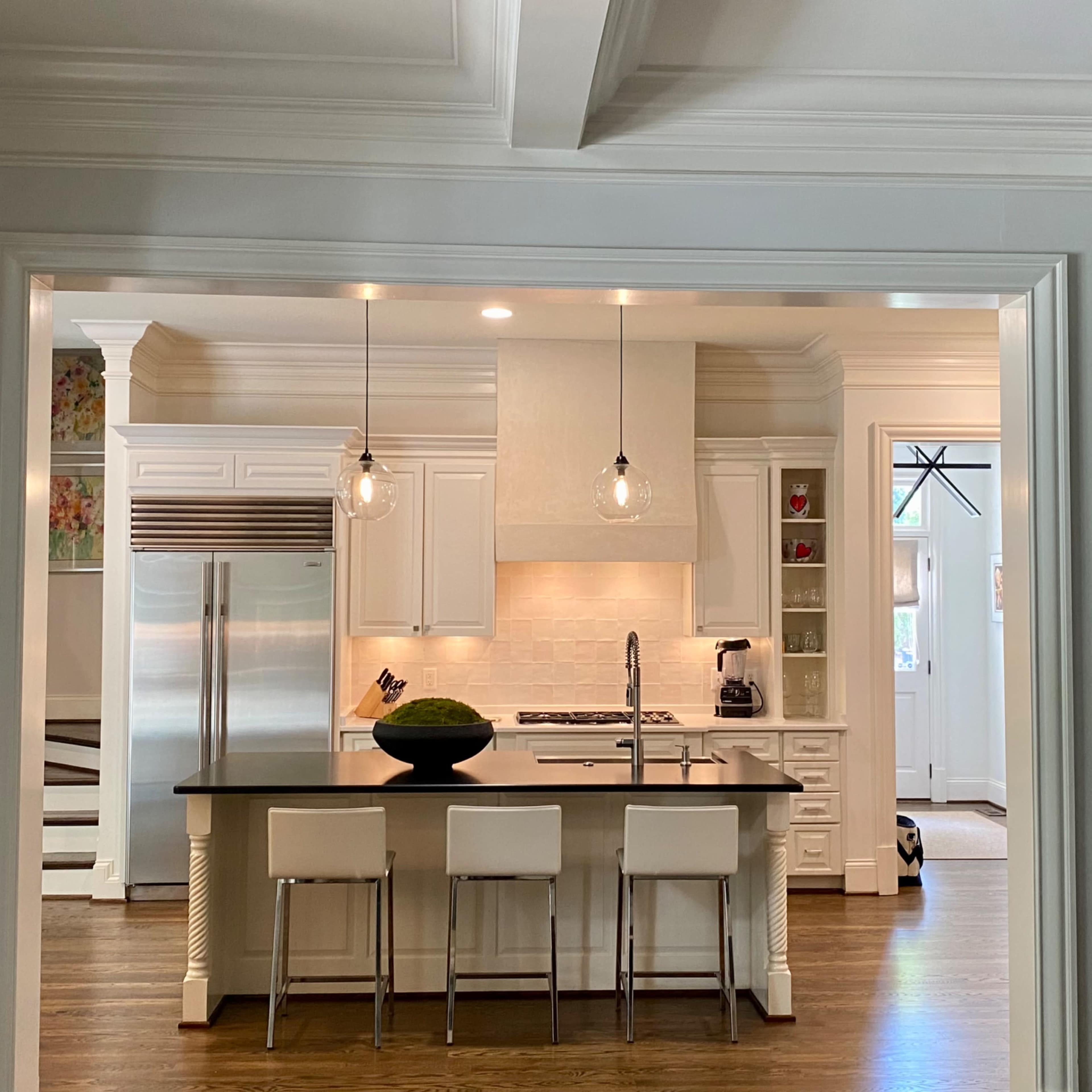 A modern kitchen features a central island with three stools, stainless steel appliances, and pendant lighting.